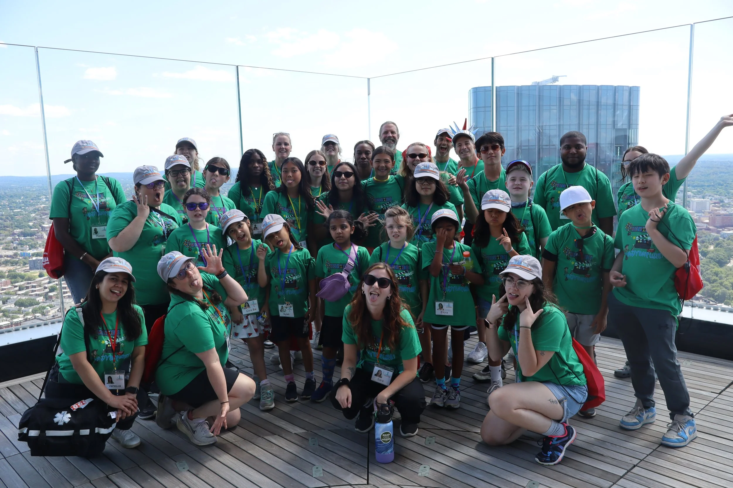 Group of children and adults on a rooftop, wearing matching green shirts with a logo, some with hats and sunglasses, posing for a photo with cityscape in the background.