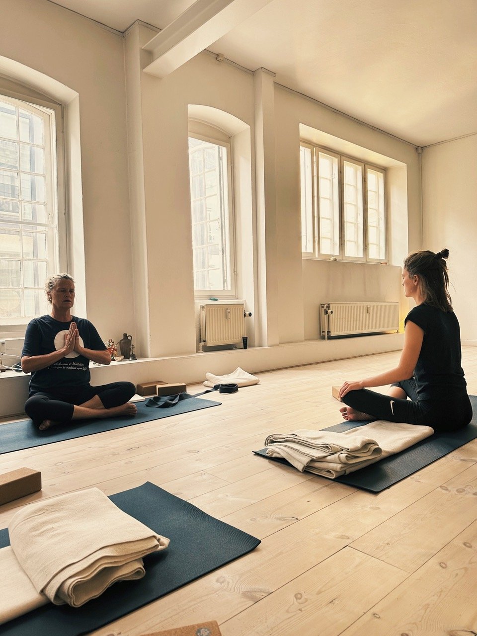 To personer der laver yoga i et lyst rum med store vinduer. De sidder på yogamåtter med tæpper og blokke på gulvet. Den ene person holder hænderne i en meditationsstilling.
