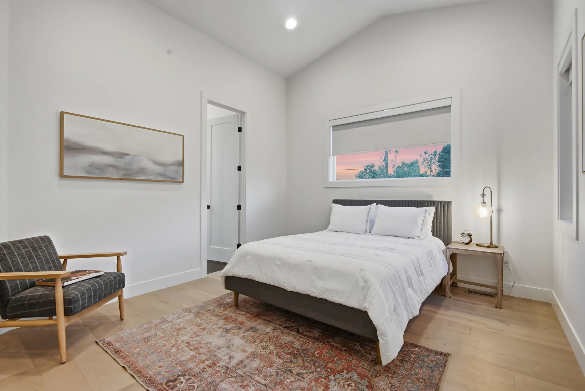 A minimalist bedroom with a white bed, a wooden side table with a lamp and clock, a mid-century modern chair with a magazine, a painting on the wall, a window showing sunset outside, and light wooden flooring.