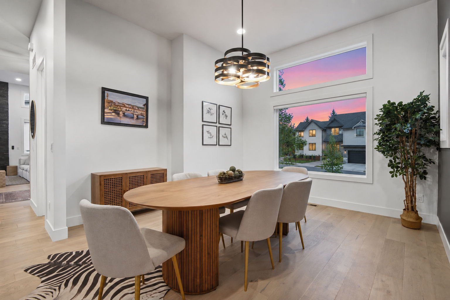 A modern dining room with an oval wooden table surrounded by six beige upholstered chairs, a black and gold pendant light fixture, large windows showing a sunset sky and houses outside, a framed picture of a cityscape, and a tall potted plant in the 