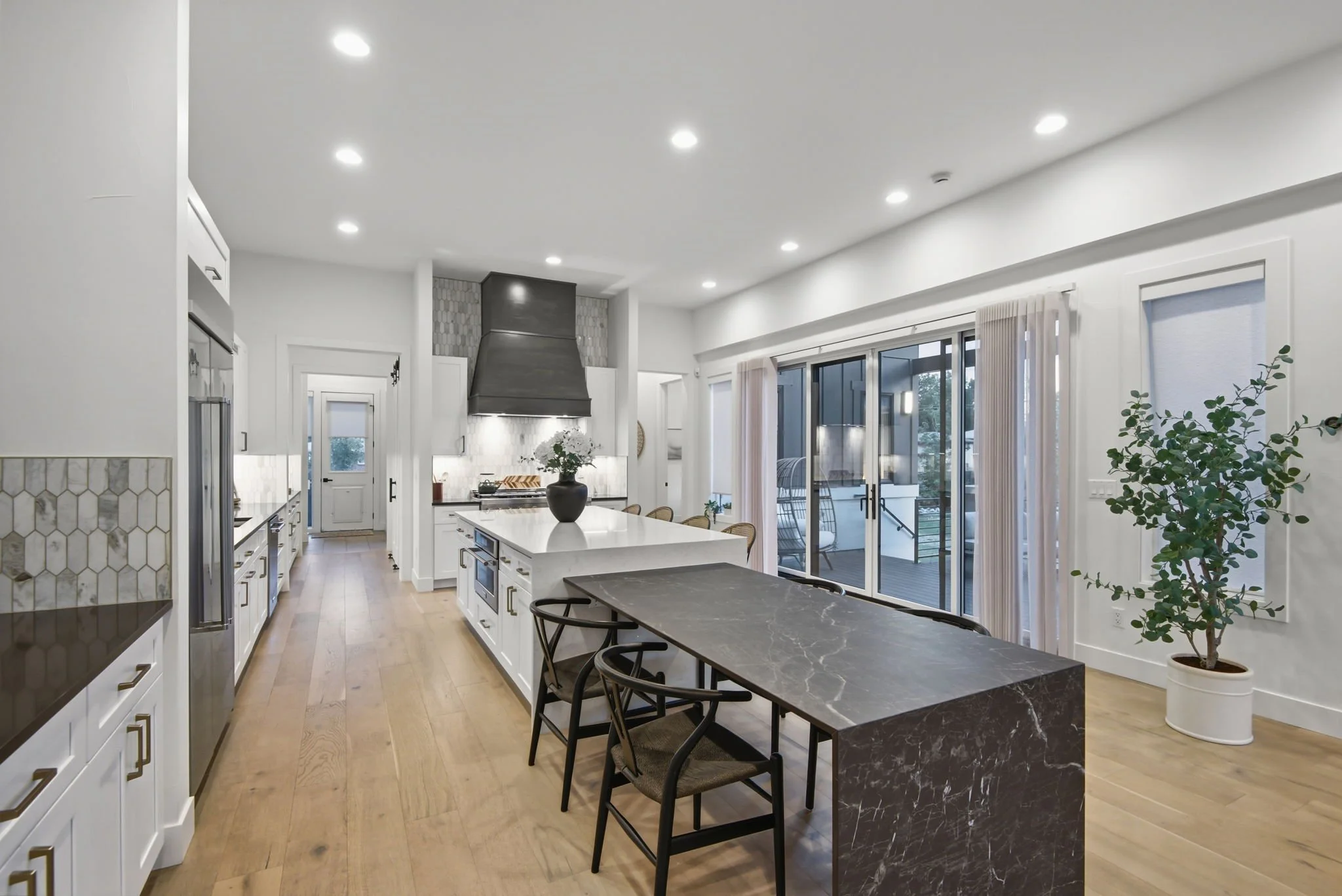 Modern kitchen with white cabinets, stainless steel appliances, a large island with a black marble countertop, a black range hood, and sliding glass doors leading to an outdoor patio, with a potted plant nearby.