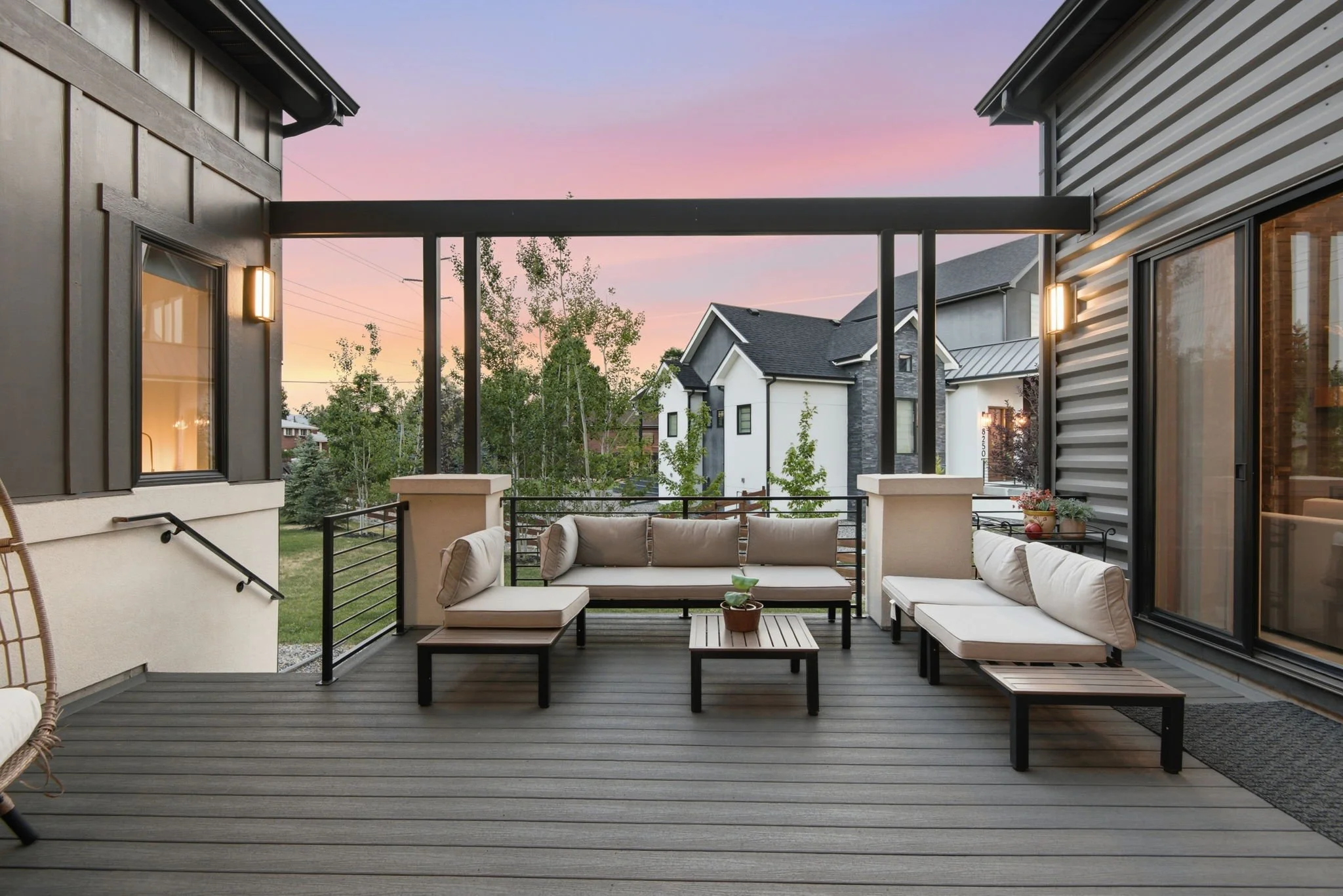 Outdoor patio with beige cushioned seating, a small wooden coffee table with a potted plant, and a view of neighboring houses and a colorful sunset sky.