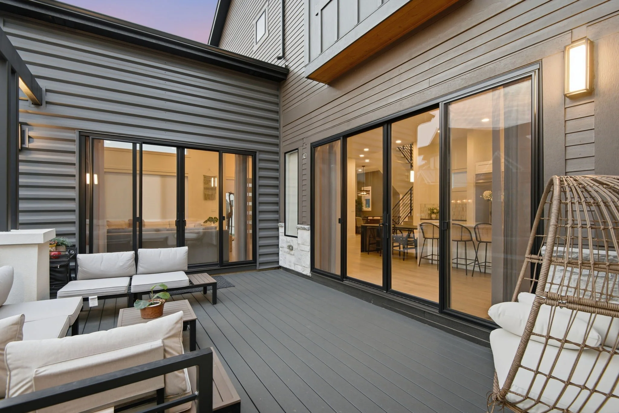 A modern patio with sliding glass doors leading into a house, outdoor seating with white cushioned chairs and a small table, and a wicker hanging chair on a gray wooden deck.
