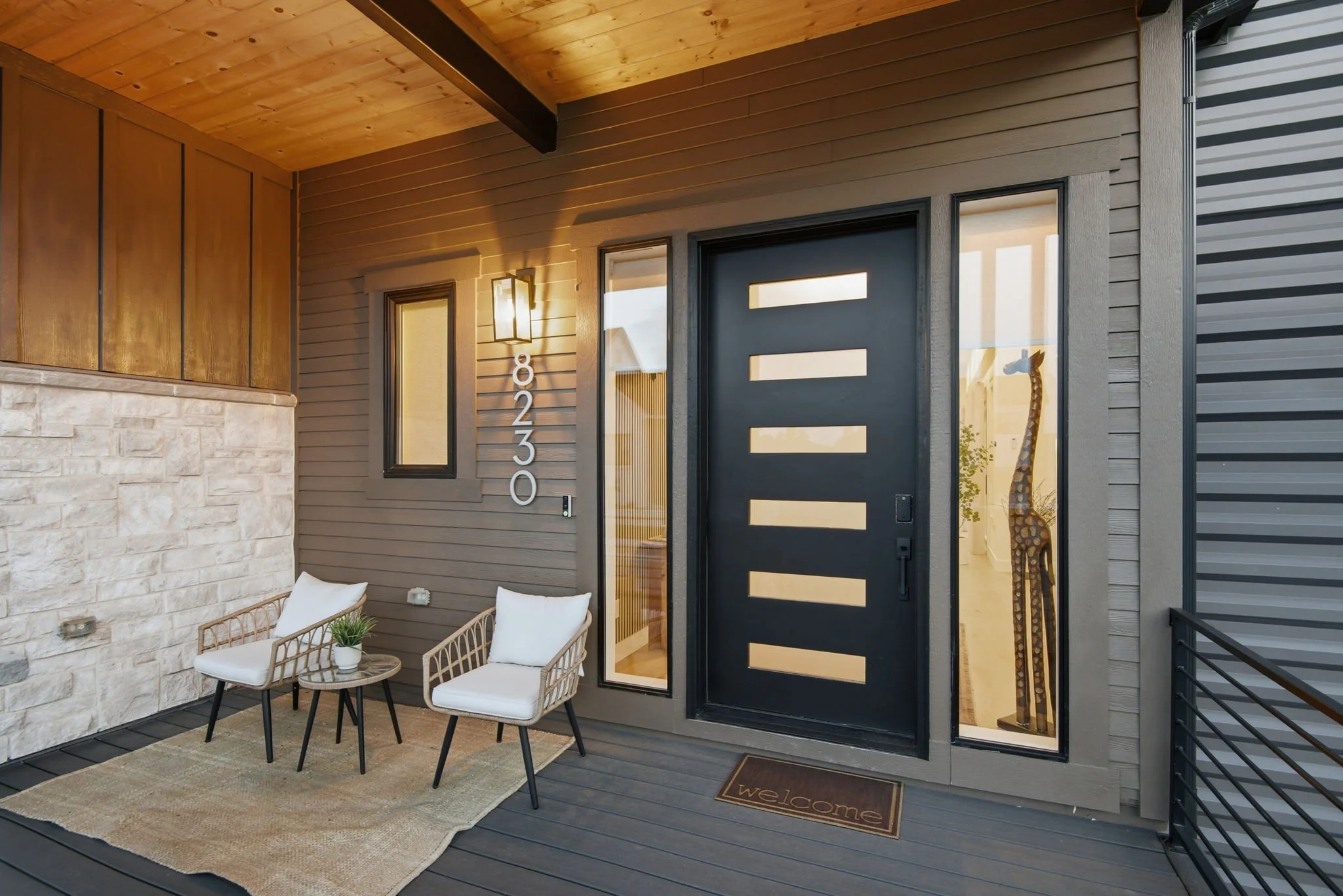 Close-up of a modern front door with vertical white stripes, flanked by a tall narrow window on the left, with a house number 8230 on the wall and a welcoming mat on the doorstep. A porch with seating and a potted plant is visible.