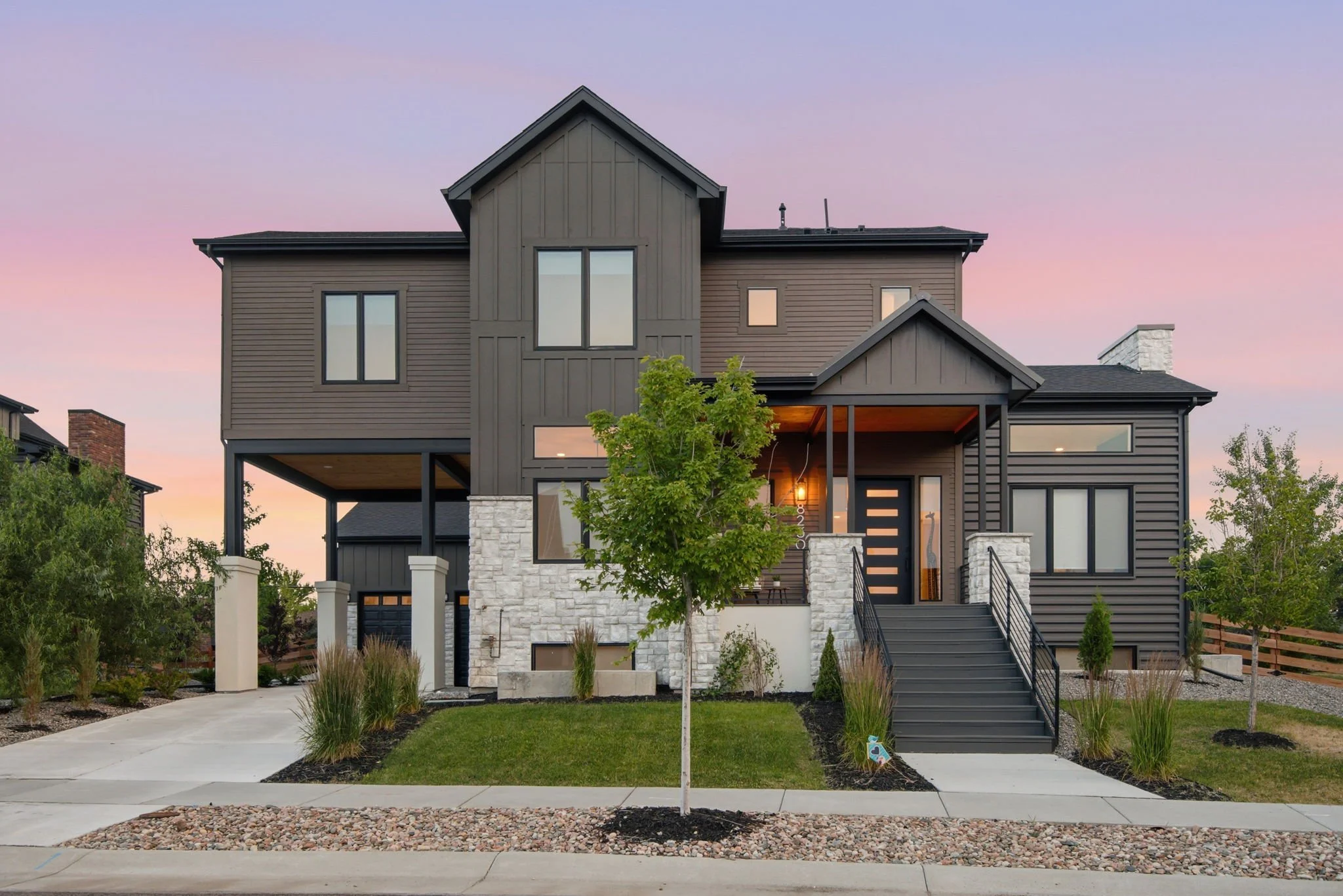 Front view of a modern, two-story house with dark gray siding, white stone accents, large windows, and stairs leading to the front entrance, surrounded by a manicured lawn and small trees, during dusk with a pink and purple sky.