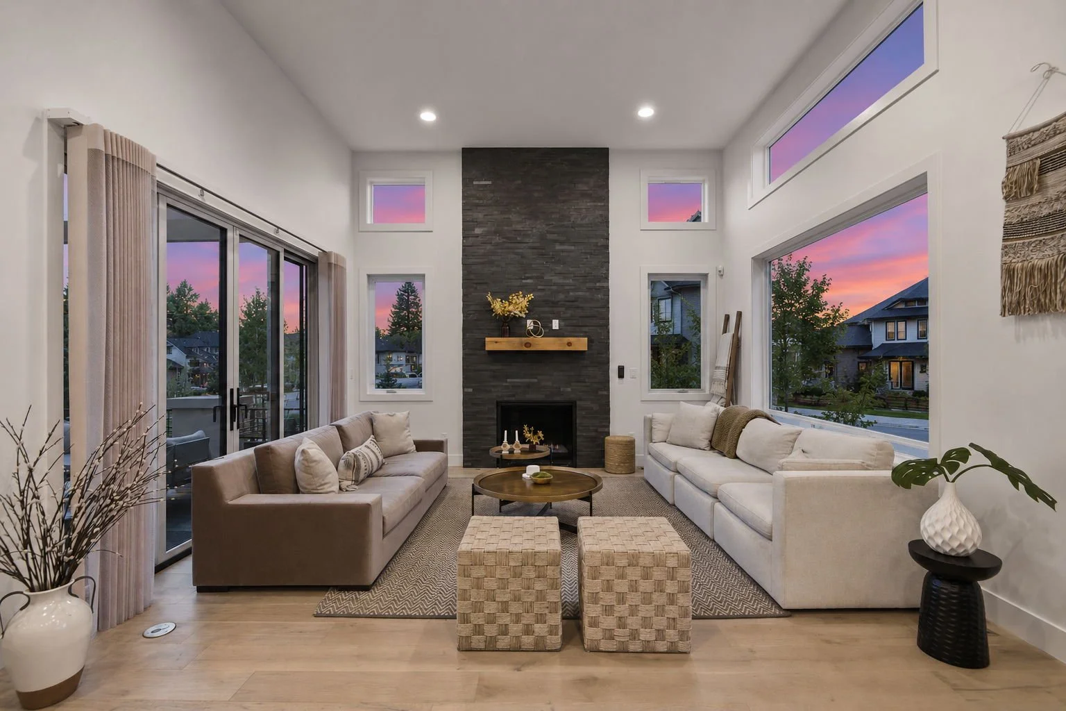 Modern living room with a fireplace, large windows showing a sunset, beige sofas, a coffee table, and decorative plants.