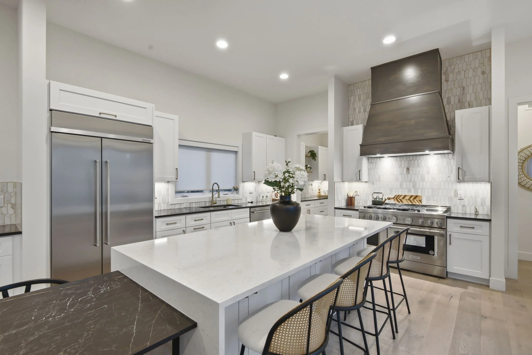 Modern kitchen with white cabinets, stainless steel appliances, a large island with a white countertop, and decorative backsplash tiles. There are chairs around the island and a black vase with white flowers on top.