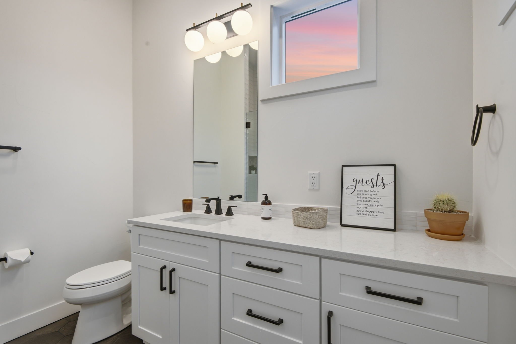 Modern bathroom with white cabinetry, a large mirror, black fixtures, a decorative framed quote, a potted cactus, a soap dispenser, a toothbrush holder, a window showing a sunset, a towel ring on the wall, and a white toilet.