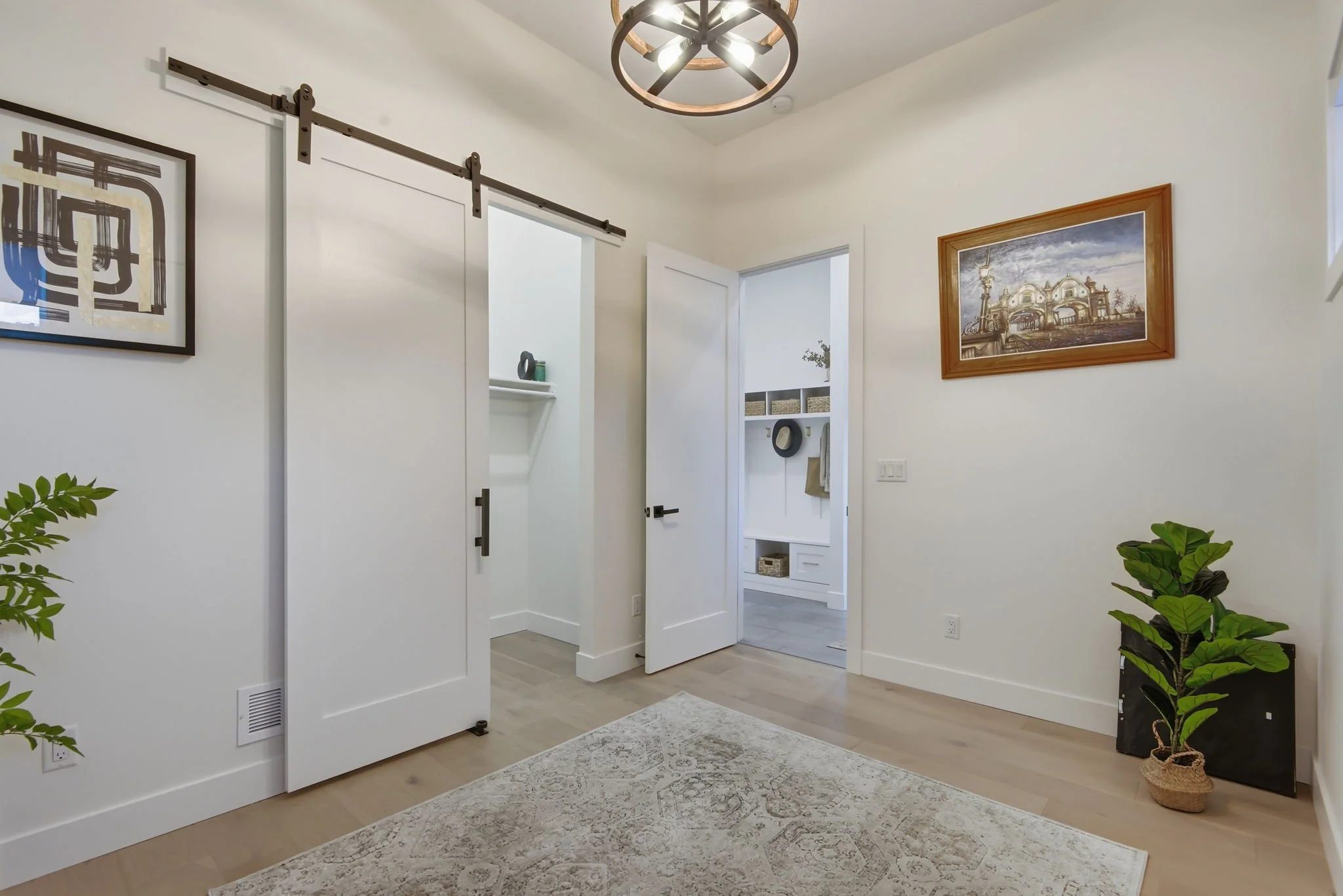 A modern entryway with white walls, a light wood floor, and decorative elements including a framed picture, a sliding barn door, a white open closet with storage baskets, a hat hanging on the wall, and a potted plant on the right side. A circular cei