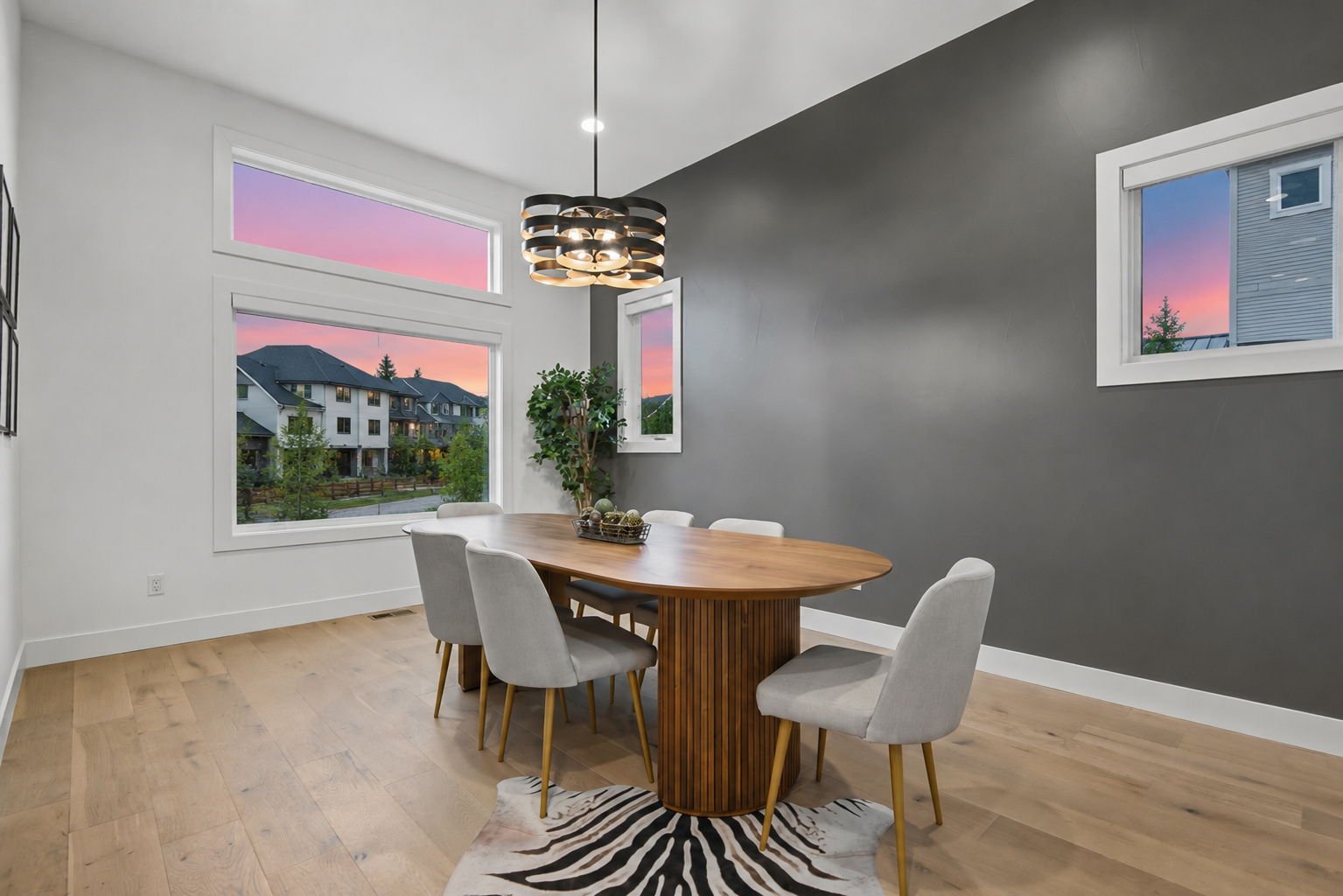 Modern dining room with a wooden table, six light gray upholstered chairs, a black and gold chandelier, large windows showing a sunset sky, a potted plant, and a zebra print rug.