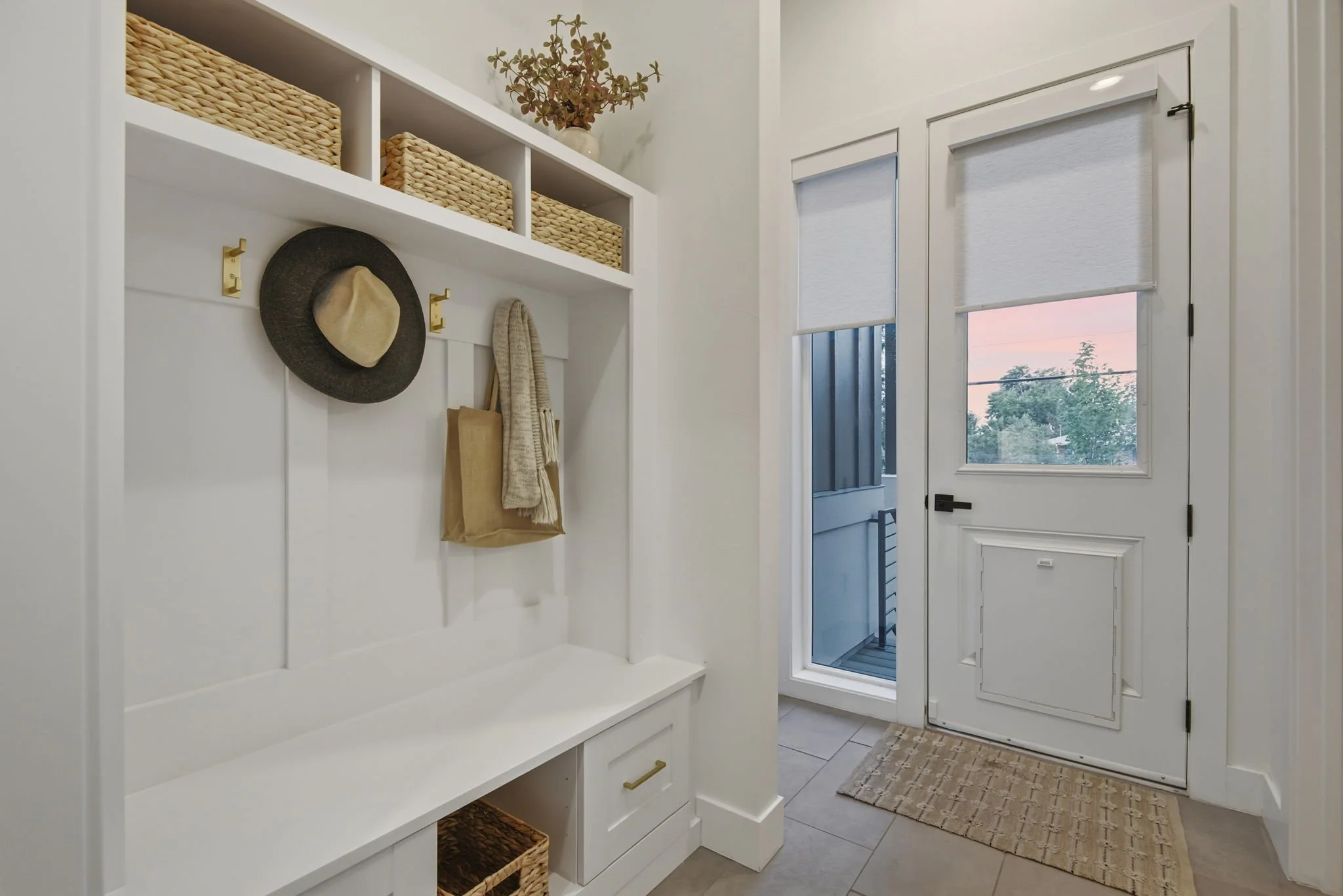 Entryway with white built-in storage unit, hooks with a hat and bag, and two windows with white blinds showing a sunset view outside.