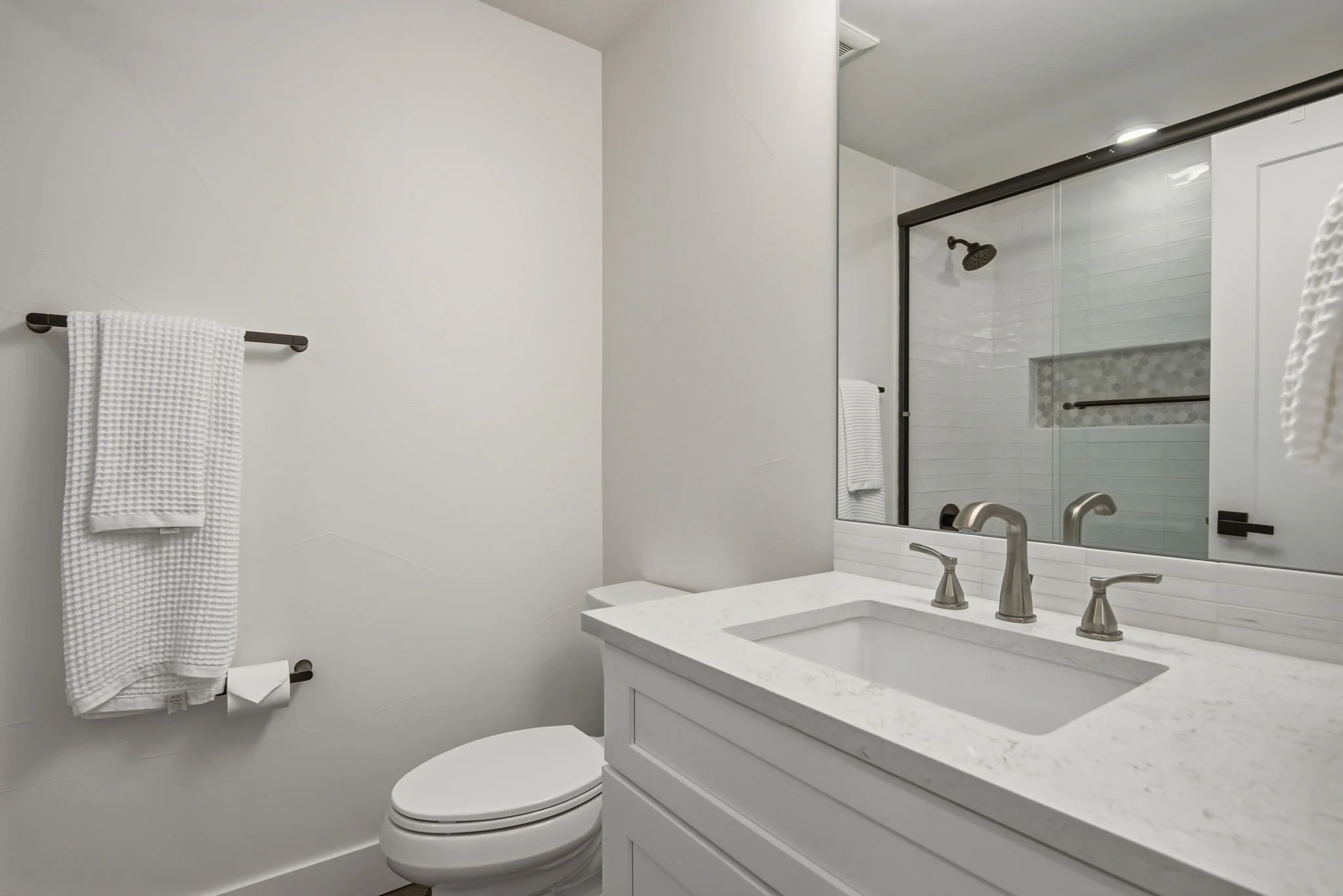 Modern bathroom with white walls, a towel hanging on a black towel rod, a white toilet, and a white vanity with a marble countertop and undermount sink. There is a large mirror above the vanity and a glass-enclosed shower with a black shower head and