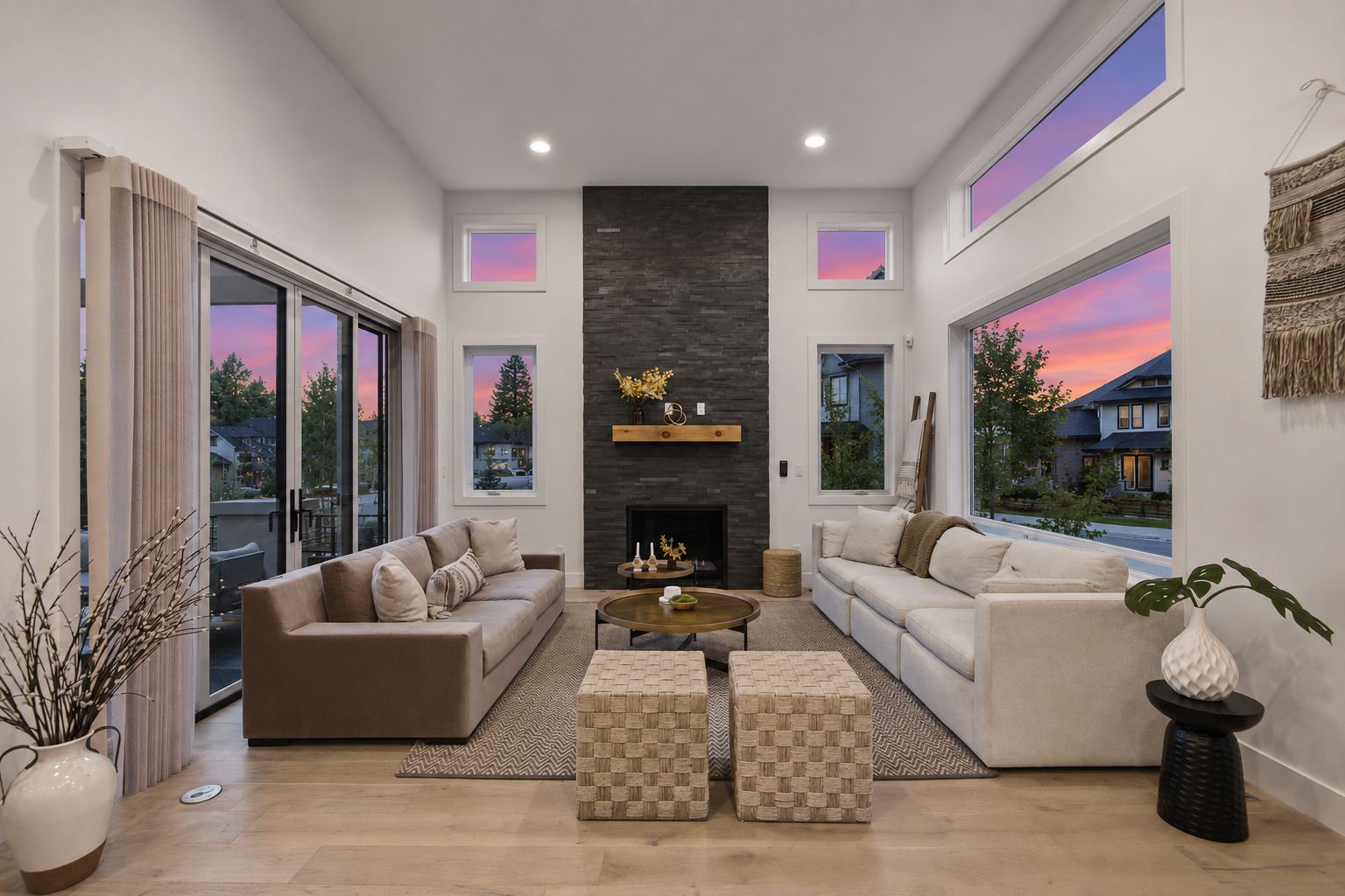 Living room with large windows, two sofas, a stone fireplace, and sunset outside.