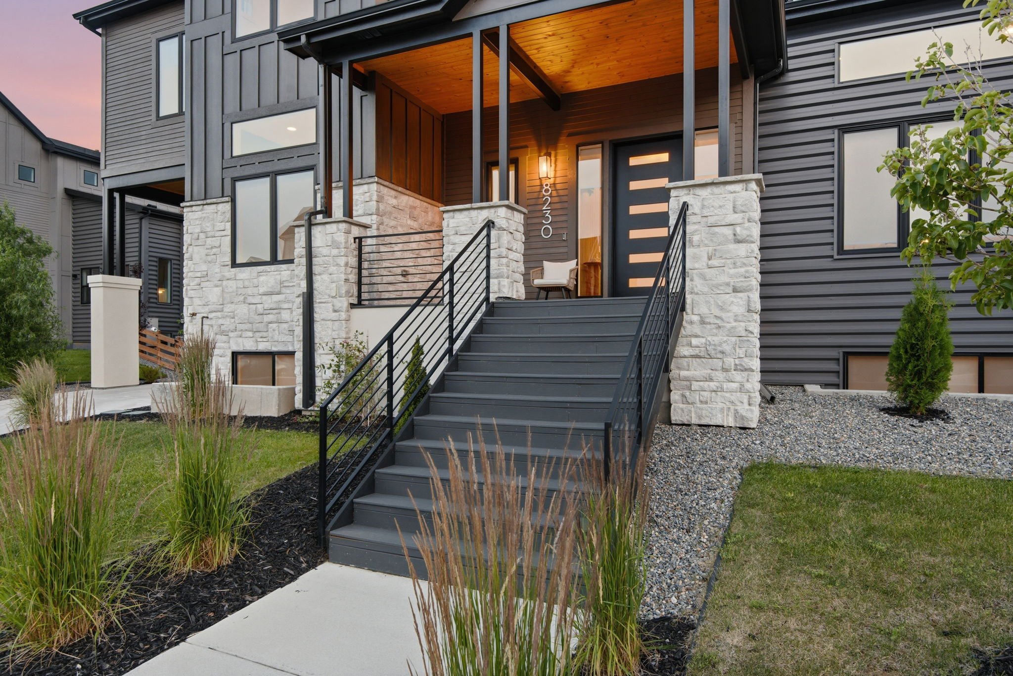 Front view of a modern townhouse with a black metal stairway leading to the entrance. The house has dark gray siding, a wooden ceiling over the porch, and stone accents on the columns. The house number 8230 is visible next to the door. Landscaping in