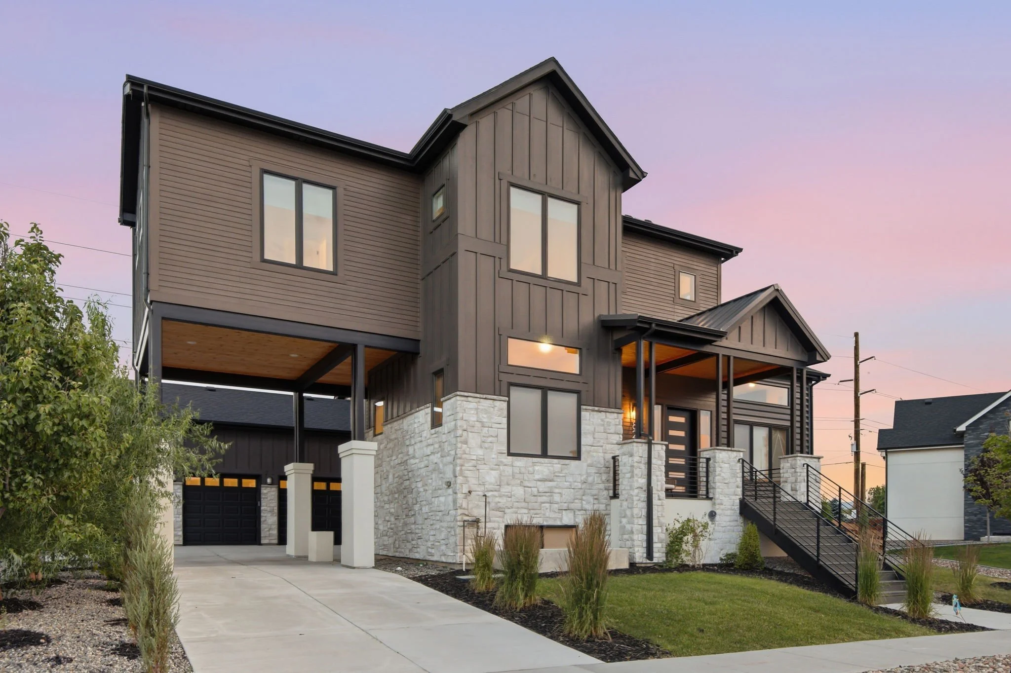 Modern two-story house with a combination of wood siding and stone exterior, large windows, and a wooden staircase leading to an entrance. Front yard has landscaping with small plants and a sidewalk.