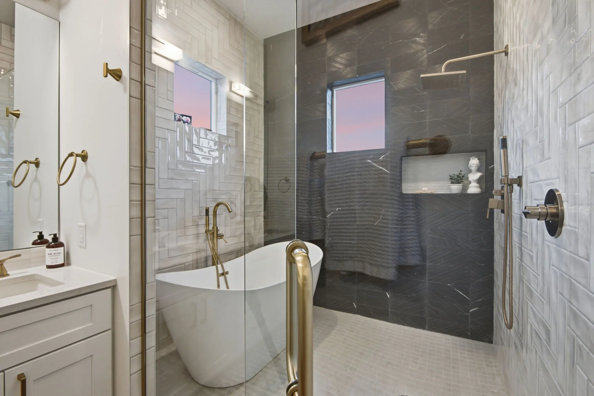 Modern bathroom with white and dark gray tiled walls, a freestanding bathtub with gold fixtures, a walk-in shower with a rainfall showerhead, a window showing a pink sky, and decorative elements like a small sculpture and potted plant.