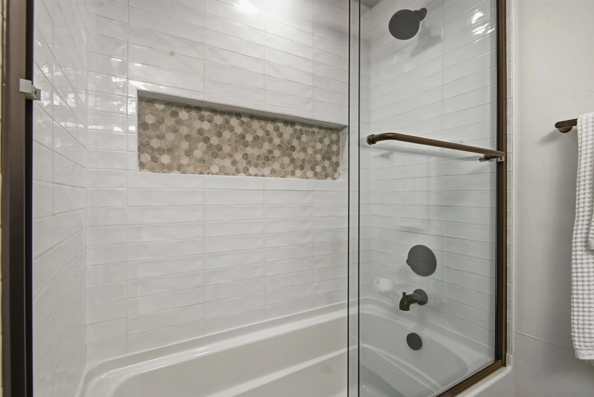 Inside a modern shower with rectangular white tiles, a built-in niche with hexagonal mosaic tiles, a glass door, a black showerhead, and a black shower control, with a white towel hanging on a towel bar.