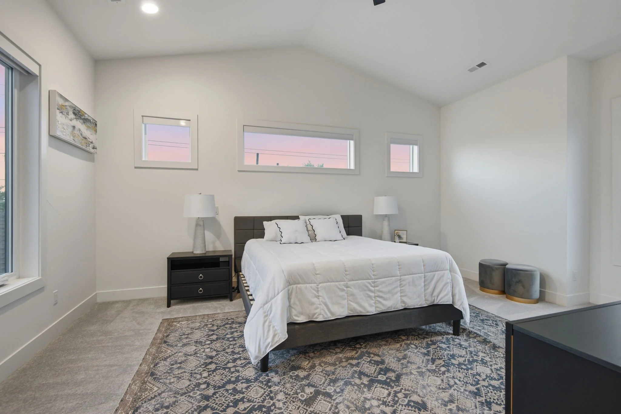 A bedroom with white walls and a high, vaulted ceiling. There are three horizontal windows above the bed, showing a pink and purple sunset sky. The bed has white bedding with black trim and a gray headboard. On each side of the bed, there are white l