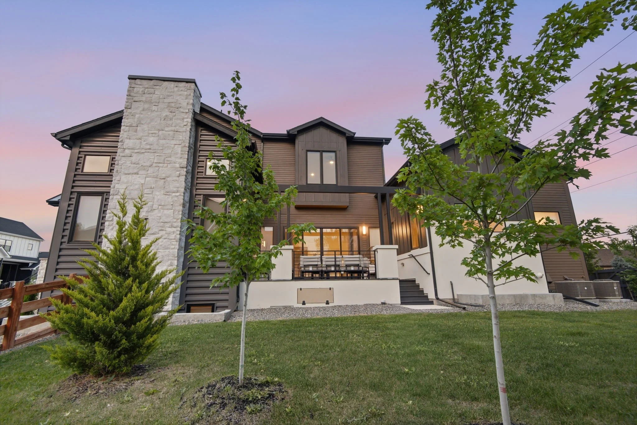 A modern house with wooden siding, large windows, a stone chimney, a balcony with dining furniture, and green trees in the front yard during twilight.
