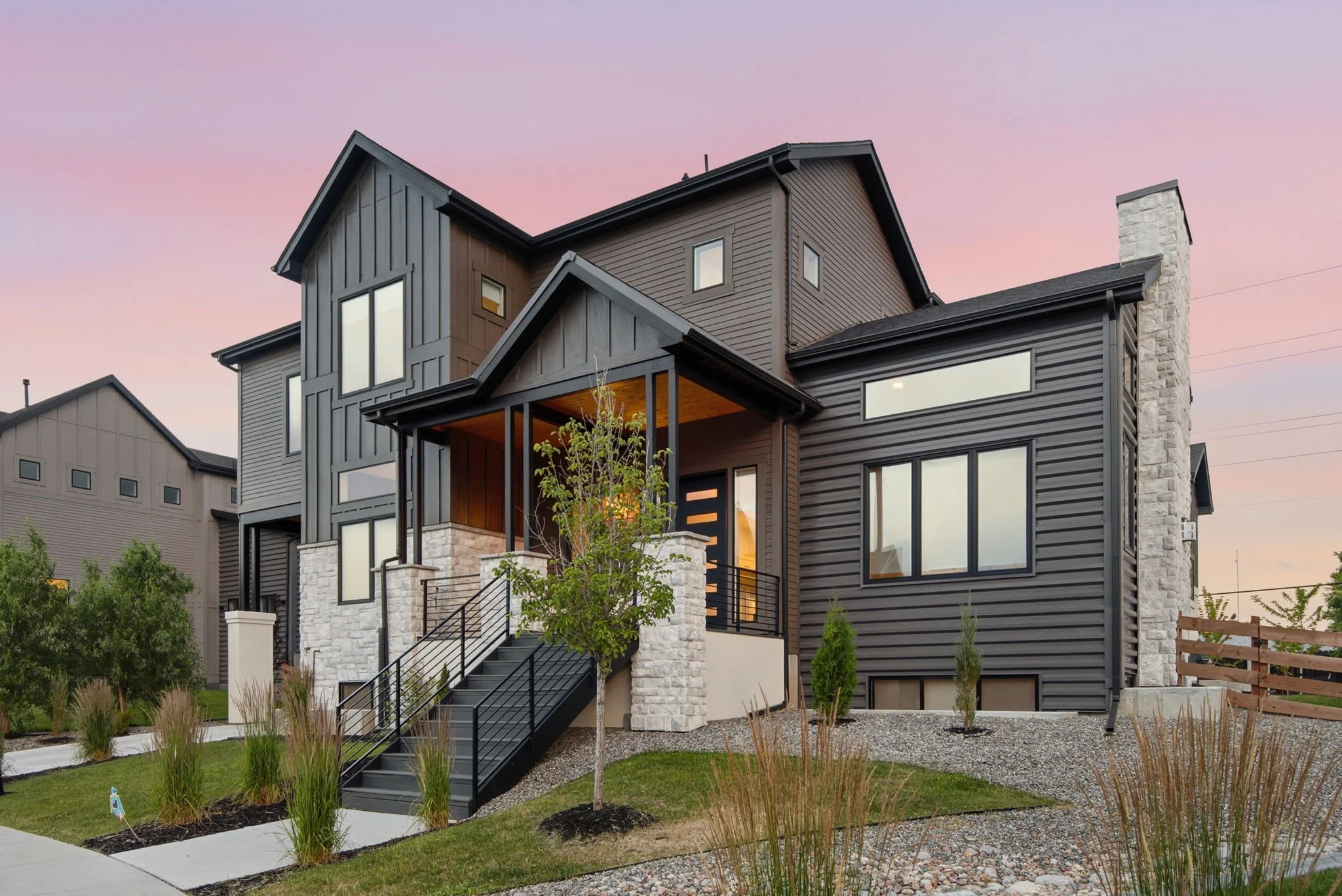 Modern multi-story house with dark gray siding, large windows, stone accents, and a black staircase leading to the front door, under a pink and purple sunset sky.