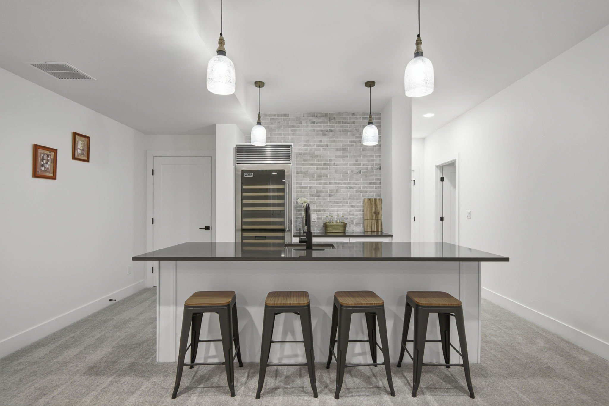 Modern kitchen with five hanging pendant lights, brick accent wall, stainless steel wine fridge, white cabinetry, and a kitchen island with four wooden stools.