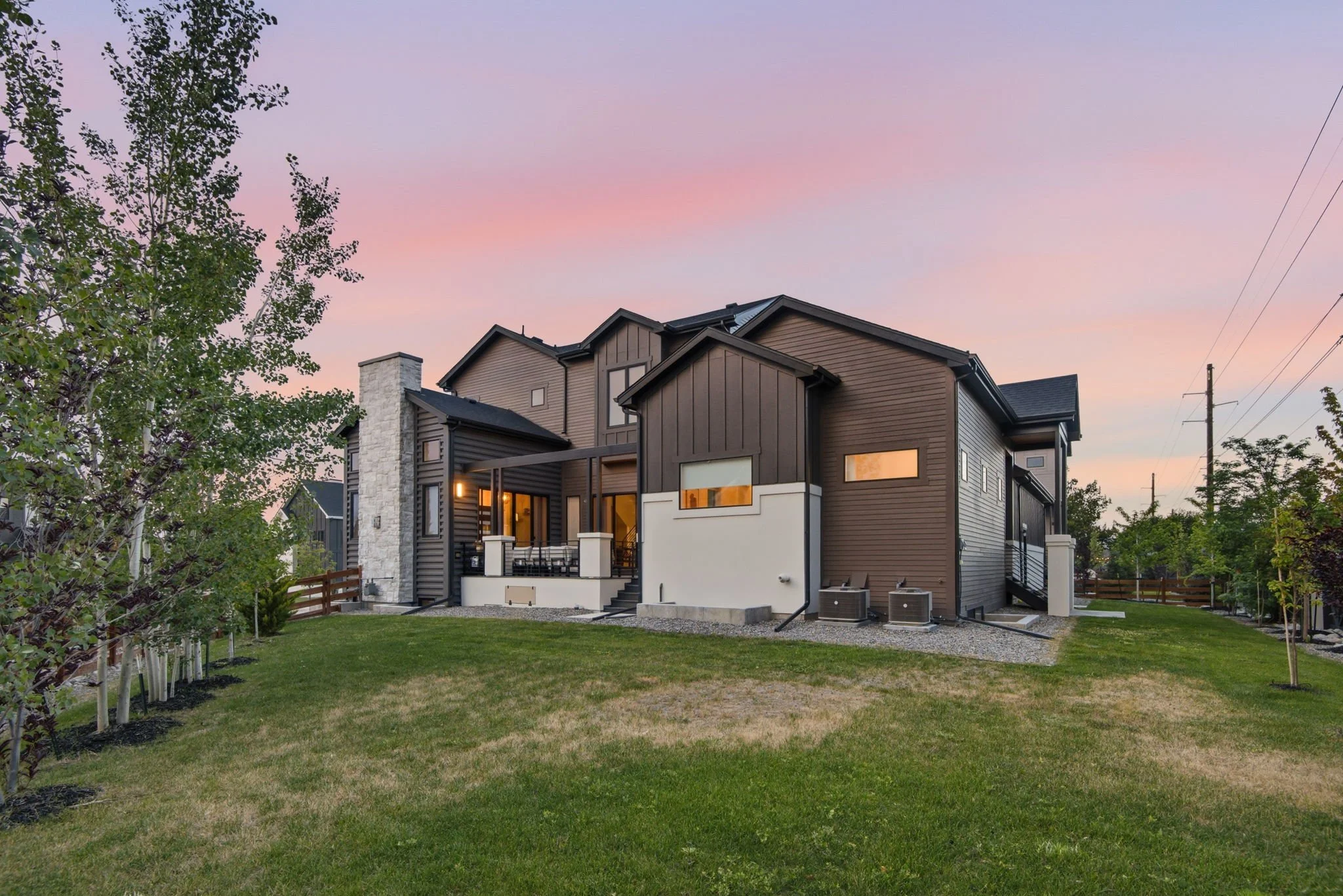 Modern two-story house with dark brown siding and a stone chimney, seen at dusk with pink and purple sky, surrounded by green lawn, trees, and some utility poles.