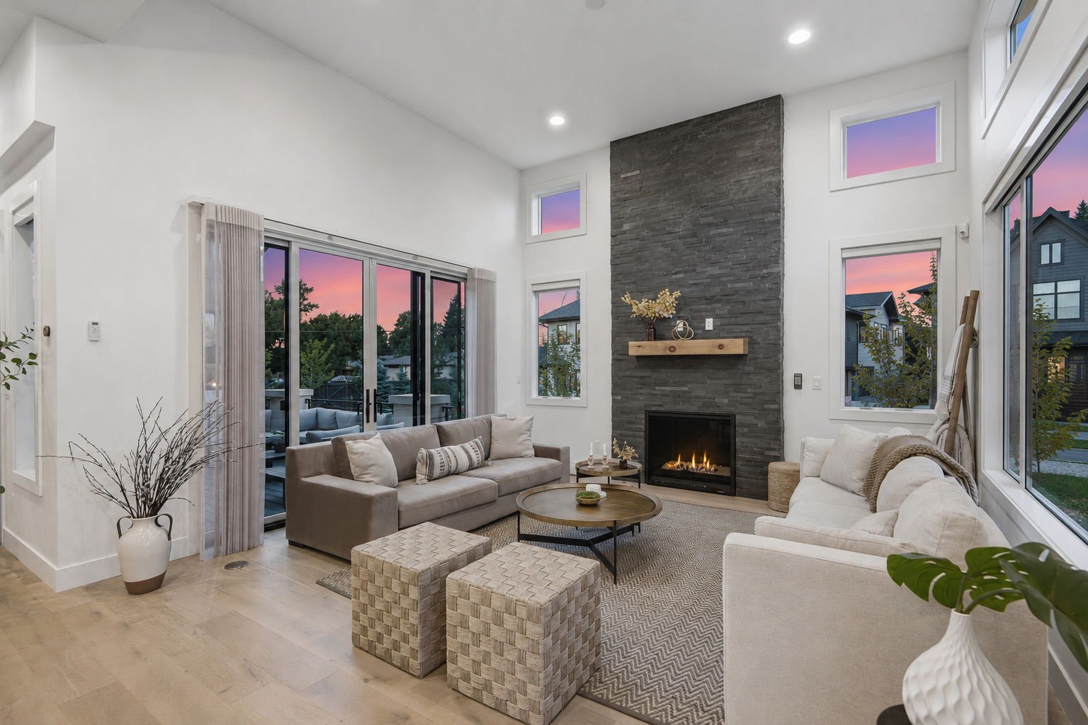 Modern living room with large windows showing sunset sky, featuring a gray fireplace, beige and white sofas, round coffee table, and decorative plants.
