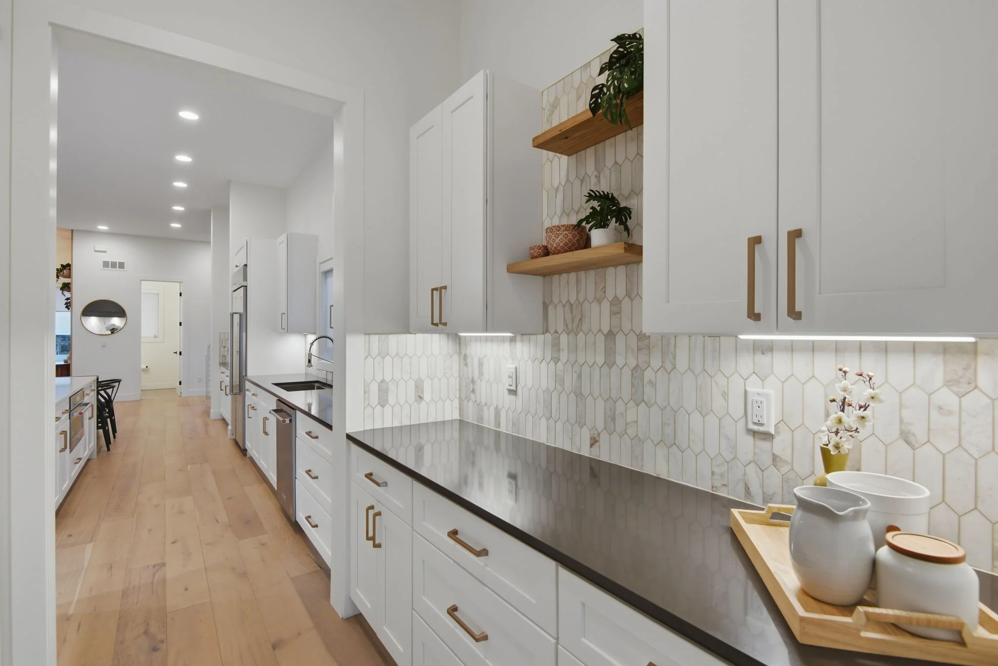 A modern white kitchen with light wood flooring, white cabinets with gold handles, and a backsplash with beige hexagonal tiles. There are open wooden shelves with plants and decor, and a dark countertop. Part of a dining area with chairs and a round 