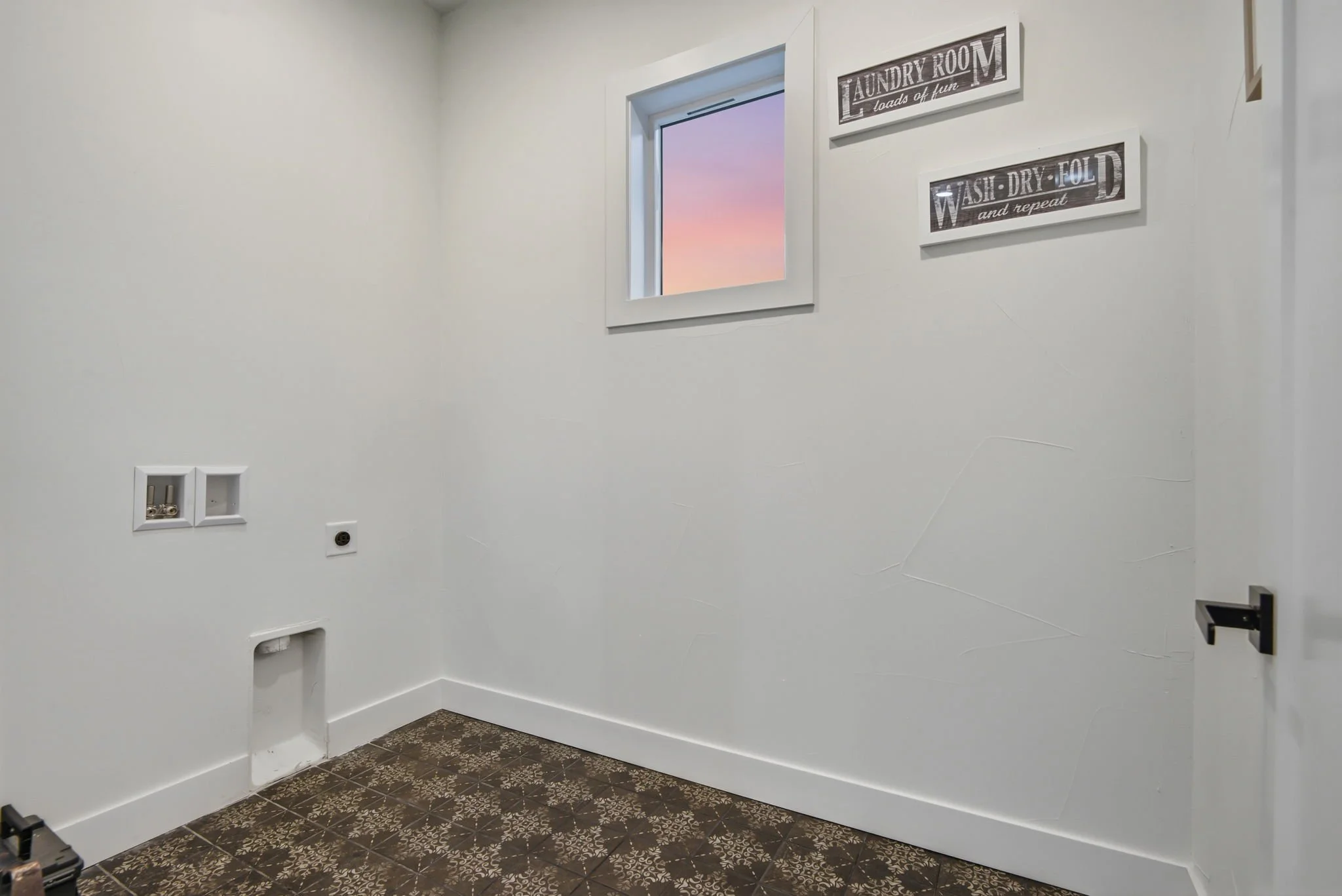 Empty laundry room with white walls, a window showing a sunset sky, and wall signs that read 'Laundry loads of fun' and 'Wash - Dry - Fold and repeat'; electrical outlets and floor tiles are visible.