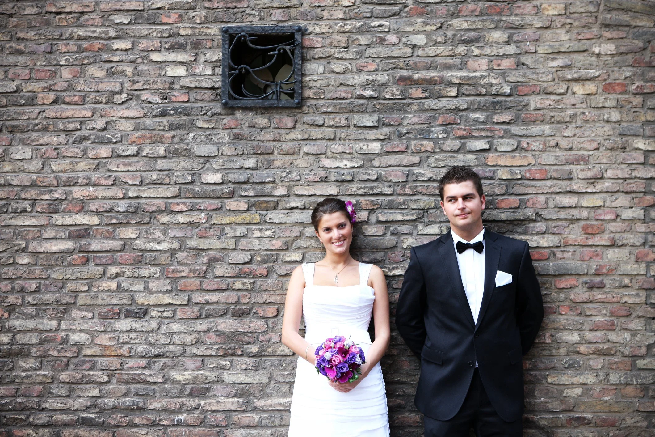 Ein Brautpaar steht vor einer Ziegelsteinmauer. Die Braut trägt ein weißes Hochzeitskleid und hält einen bunten Blumenstrauß, während der Bräutigam einen schwarzen Anzug mit Fliege trägt.