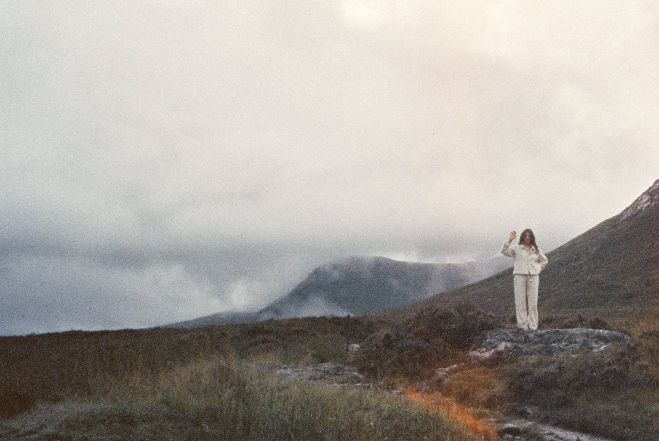A woman stands on a rock in a mountainous landscape with misty mountains in the background, waving.