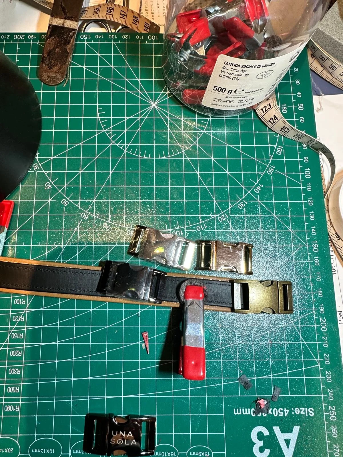 Attrezzi e materiali per il fai da te e il bricolage, tra cui un cinturino in pelle, una pinza, clip metalliche, un rotella di misurazione e una scatola di viti con testa rossa, su un tappetino da taglio verde