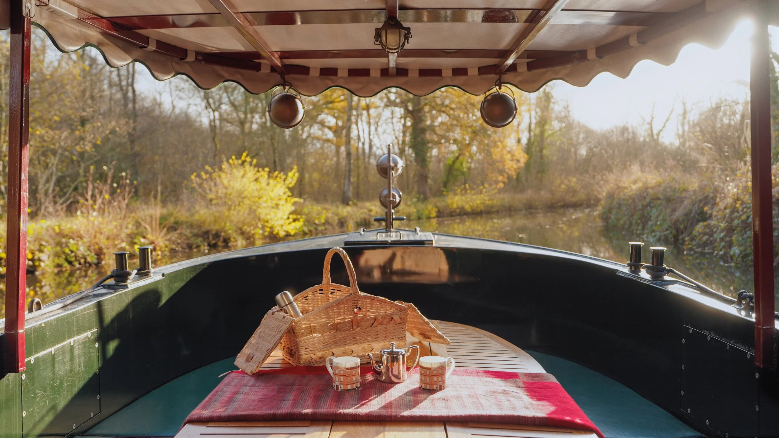 The front of a boat with a red canopy, showing a small wooden table set for tea with a red tablecloth, a wicker picnic basket, two cups, a teapot, and a metal flask. The boat is on a river with trees and autumn foliage in the background.
