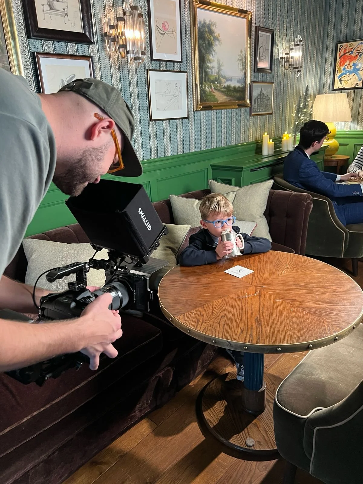 A man with a camera filming a young boy with glasses drinking from a metal mug at a wooden table in a cozy, decorated room.