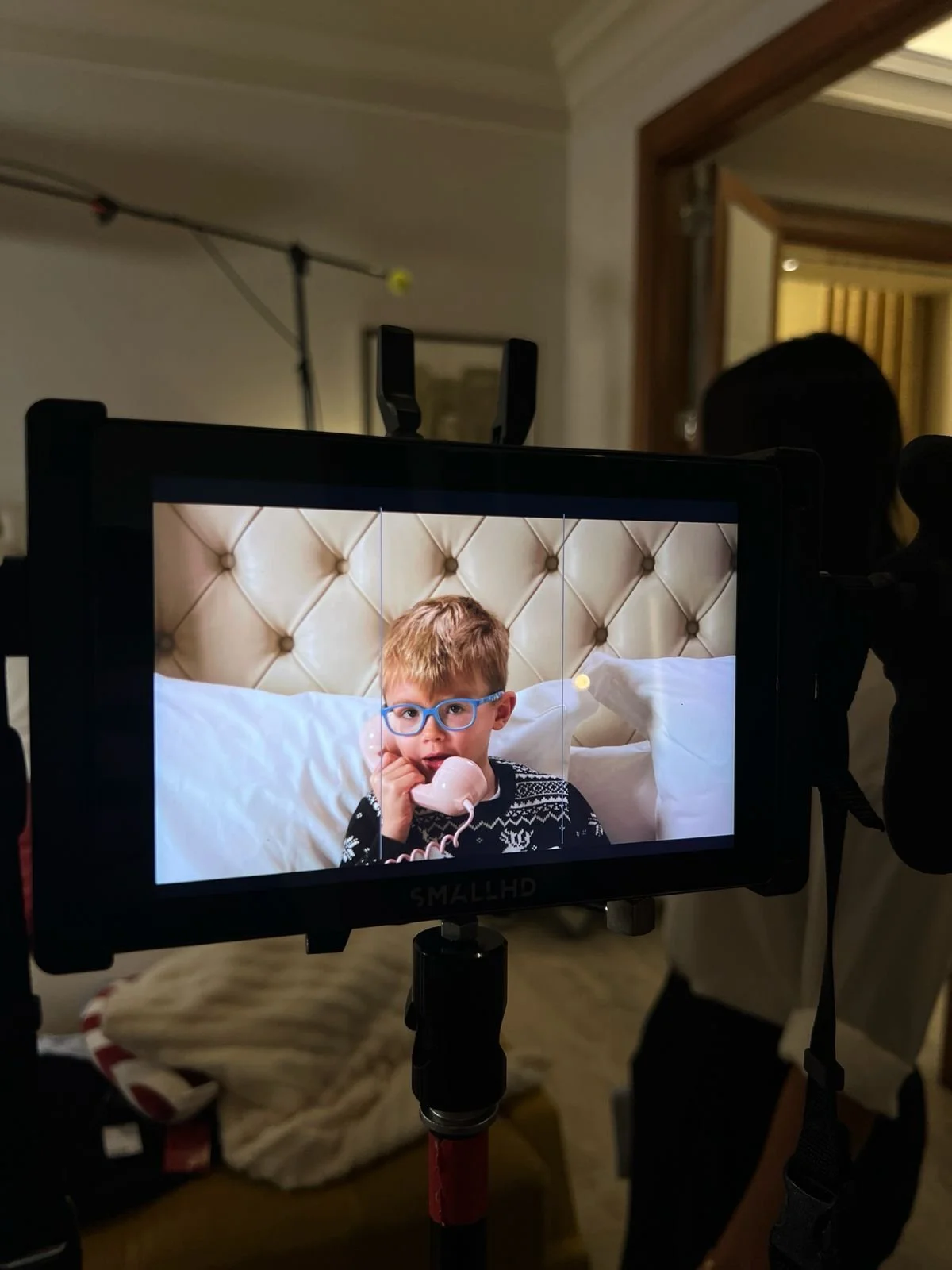 On a film or photo set, a young boy with blonde hair, blue glasses, and a patterned sweater is sitting on a bed with a beige upholstered headboard, talking on a pink phone, as seen through a camera monitor. Behind, there are people and equipment, and