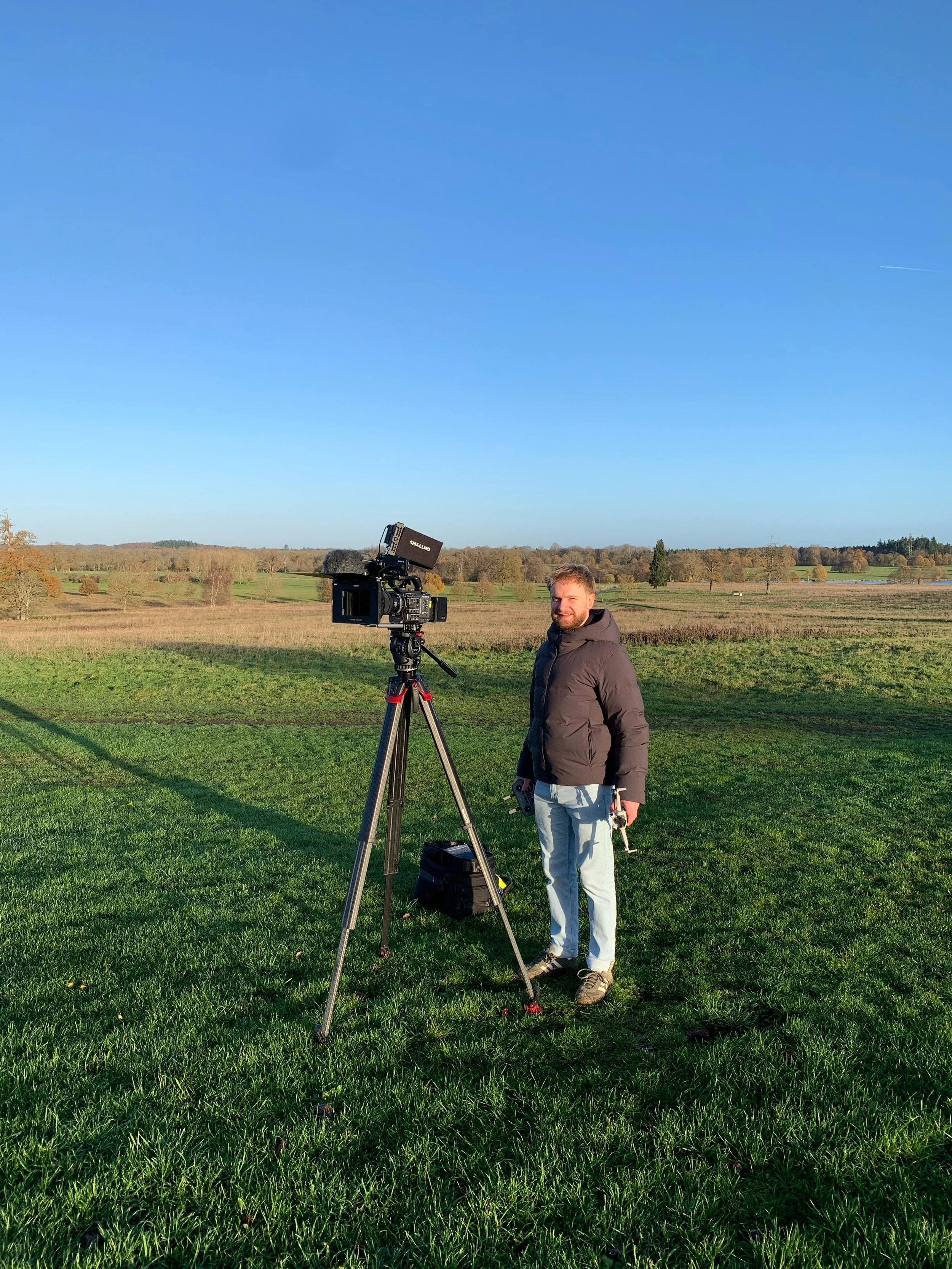 A man standing outdoors on a grassy field next to a professional video camera mounted on a tripod, with a clear blue sky and trees in the background.