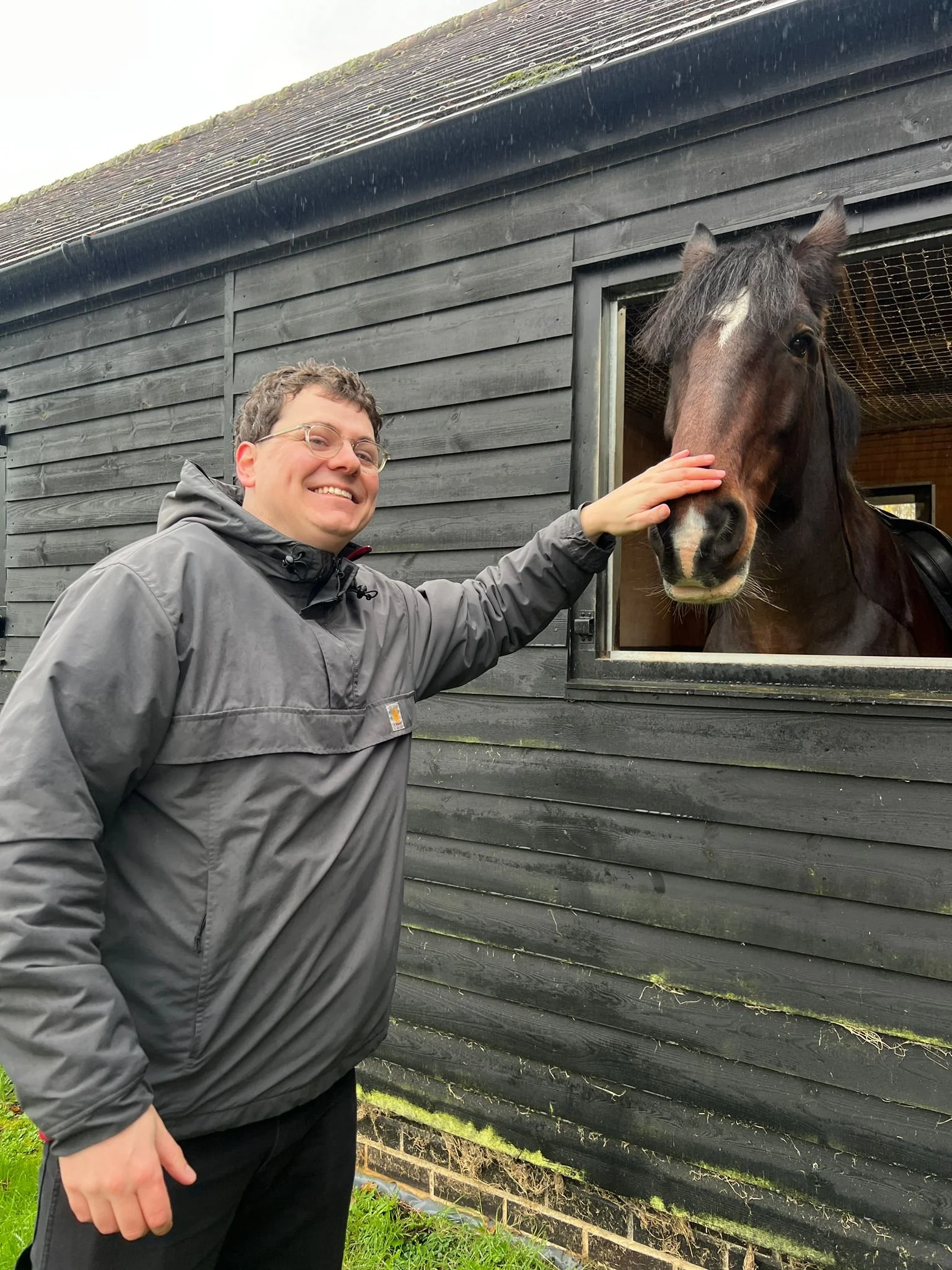 A man wearing glasses and a gray jacket petting a horse's head through a stable window, standing outdoors near a black wooden barn.