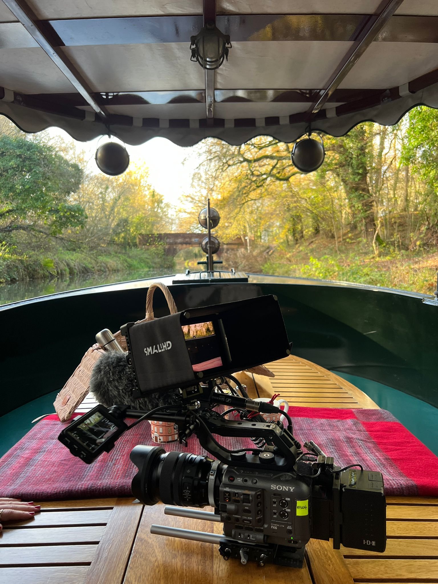 Camera and filming equipment set up on a boat with a view of a canal and trees during daylight.