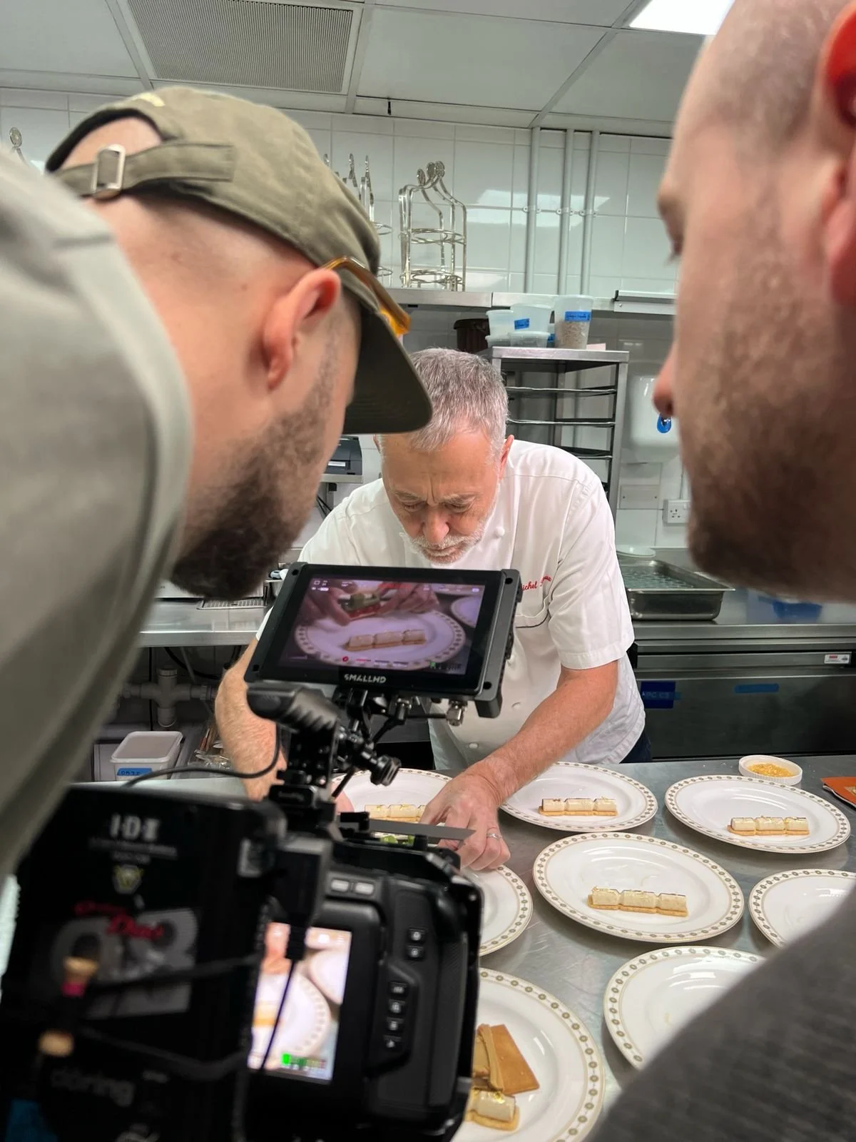 A chef is plating desserts on plates while two camera operators film him in a commercial kitchen.