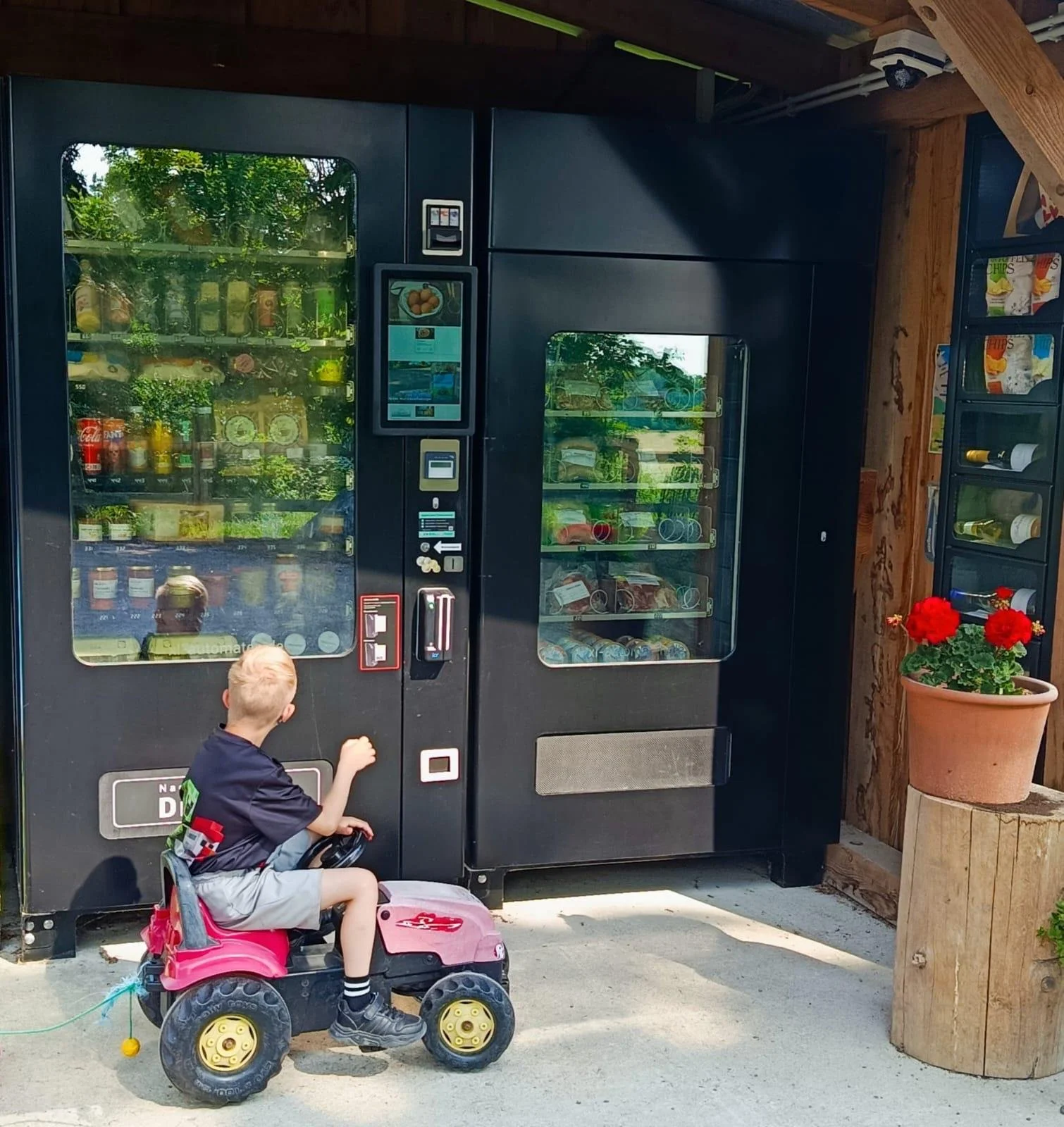Ein Junge sitzt auf einem pinken Spielzeugauto und hält eine Hand an eine Snack-Automat. Es gibt zwei Snack-Automaten, einen mit Getränken und einen mit Snacks. Es ist ein Außenbereich mit Holzverkleidung und Topfpflanzen.