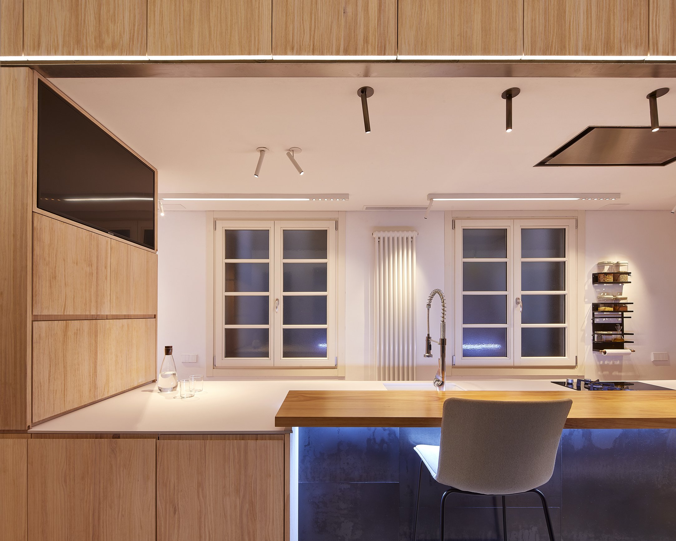 Cocina moderna con isla de madera, ventanas blancas, iluminación empotrada y estanterías organizadas.