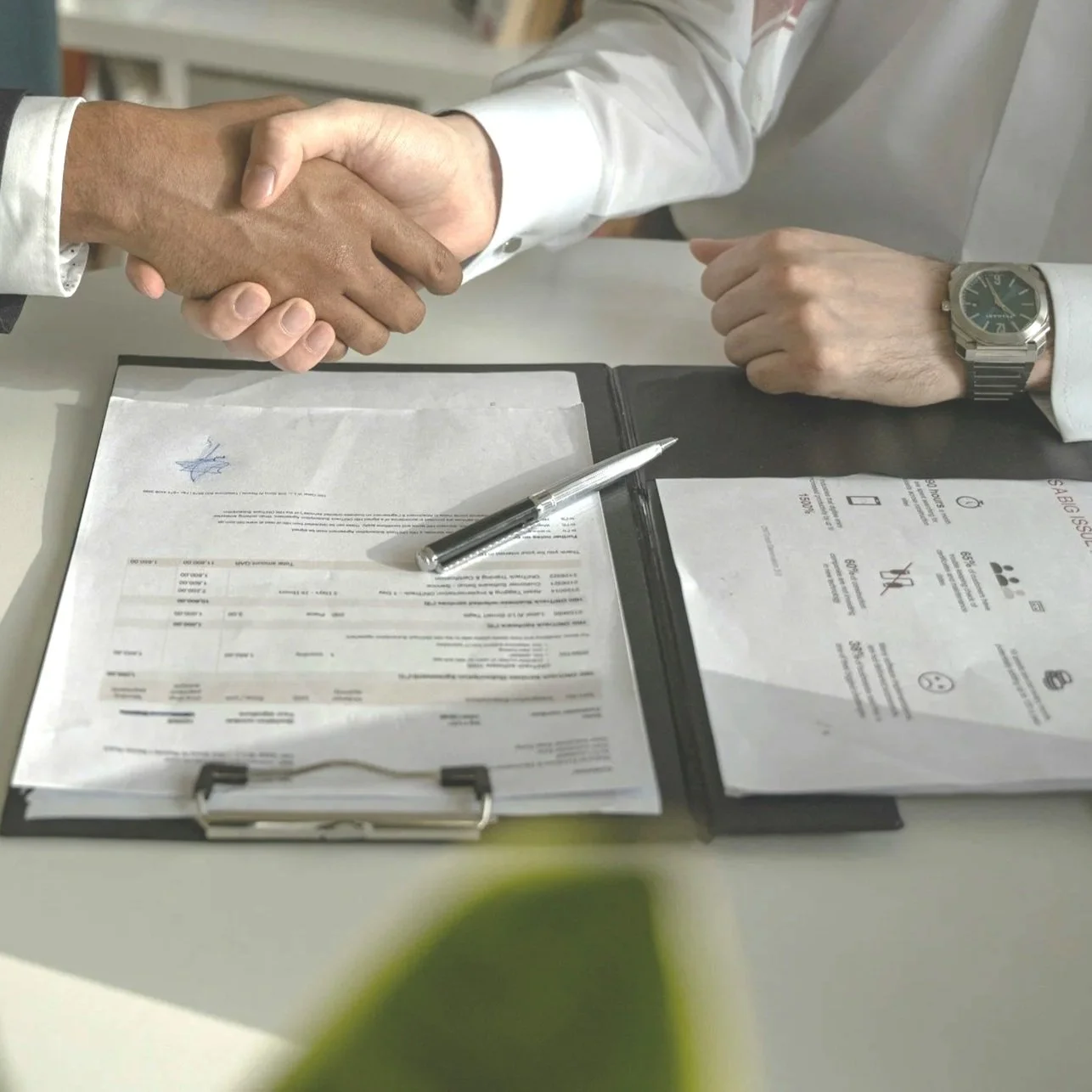 Två personer, en man och en kvinna, som sitter vid ett skrivbord i ett kontorsrum, ger varandra en high five och ler. Det finns papper, en laptop, kaffekoppar och kontorsutrustning på bordet.