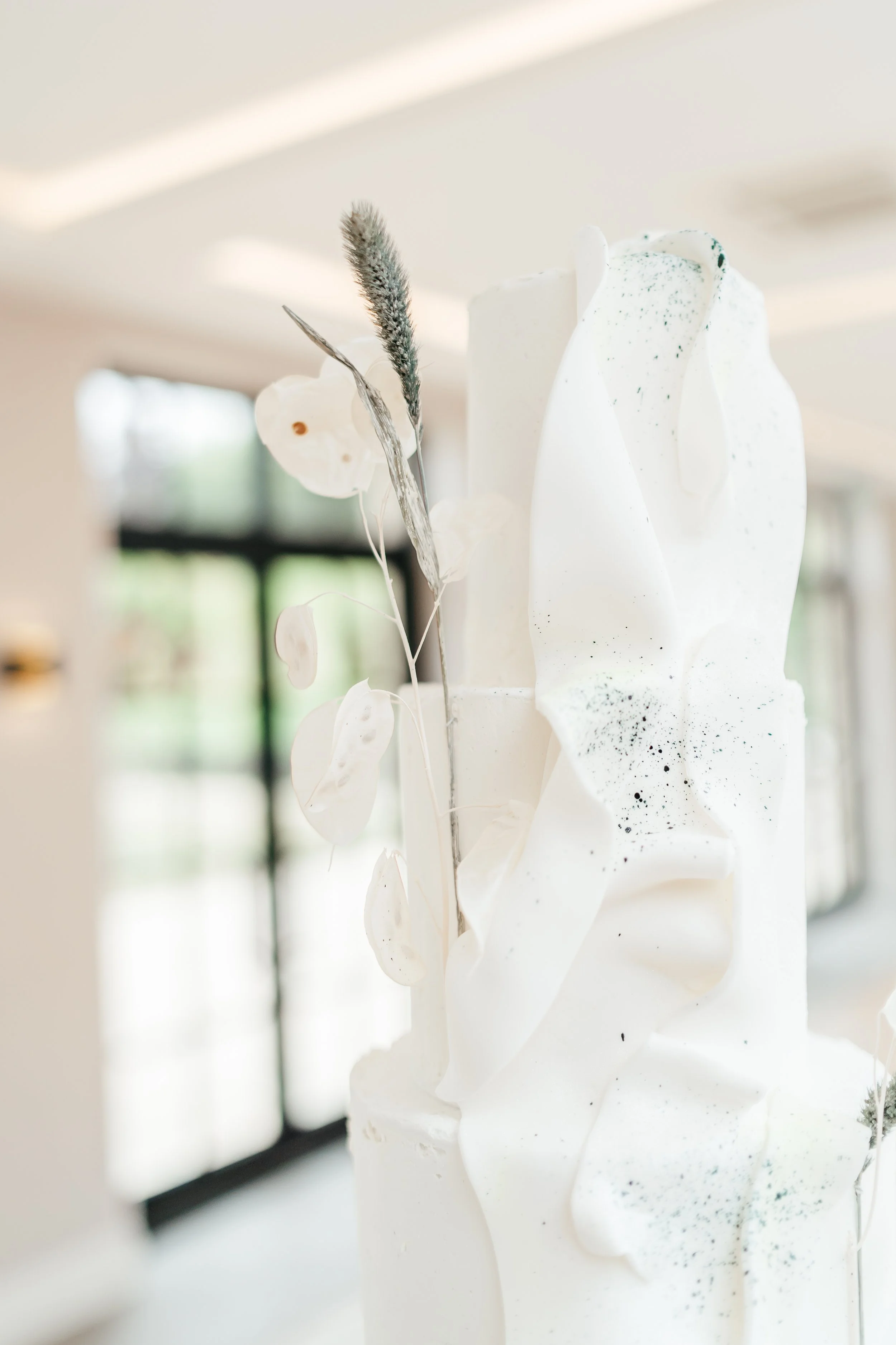 Close-up of a tall white wedding cake decorated with white and black sprigs of dried flowers and abstract white icing accents, in a bright room with large windows.