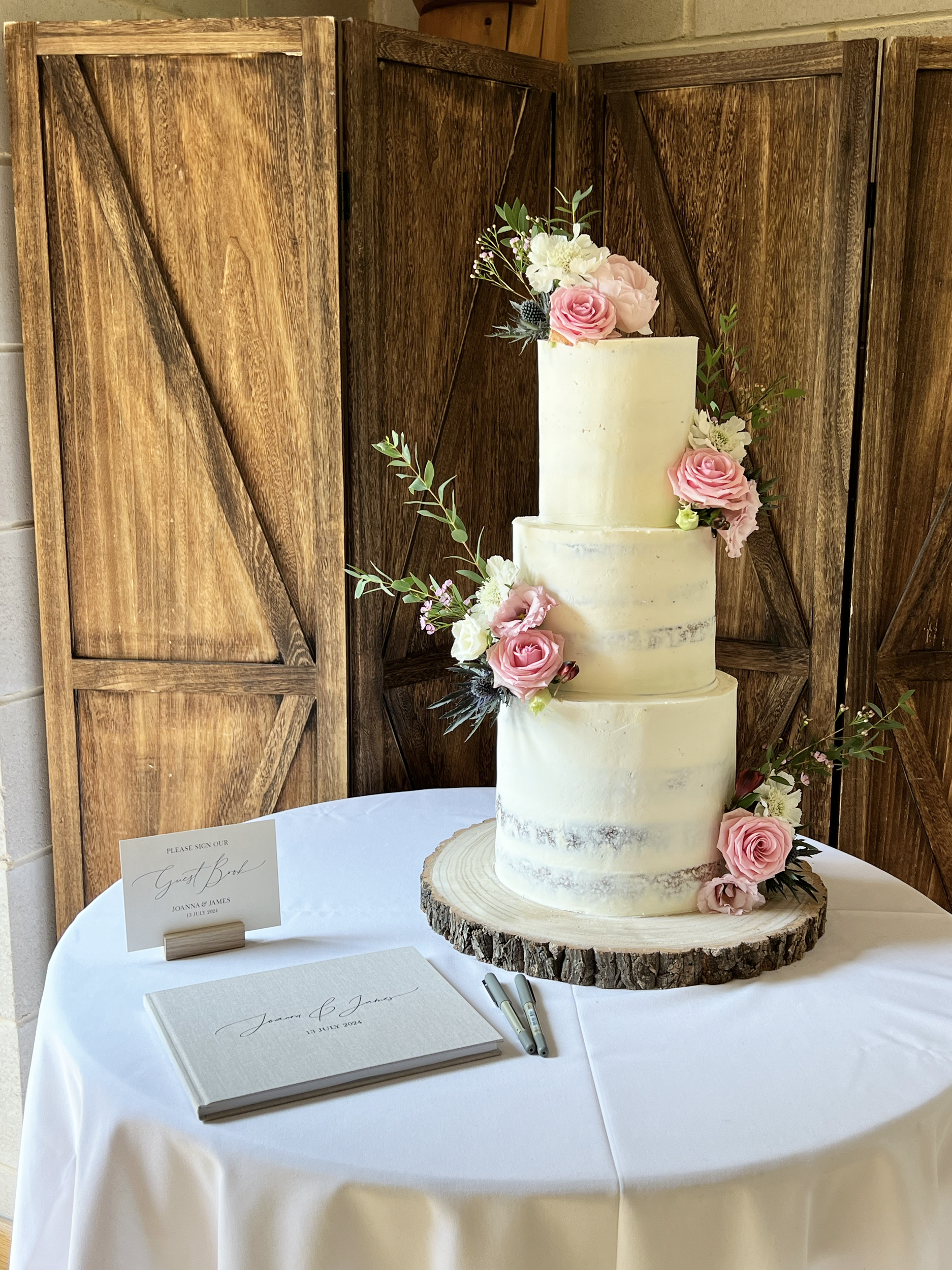 A three-tier semi-naked wedding cake with pink roses and greenery, displayed on a rustic wooden slice on a round table with a white tablecloth. A guest book and pens are placed nearby.