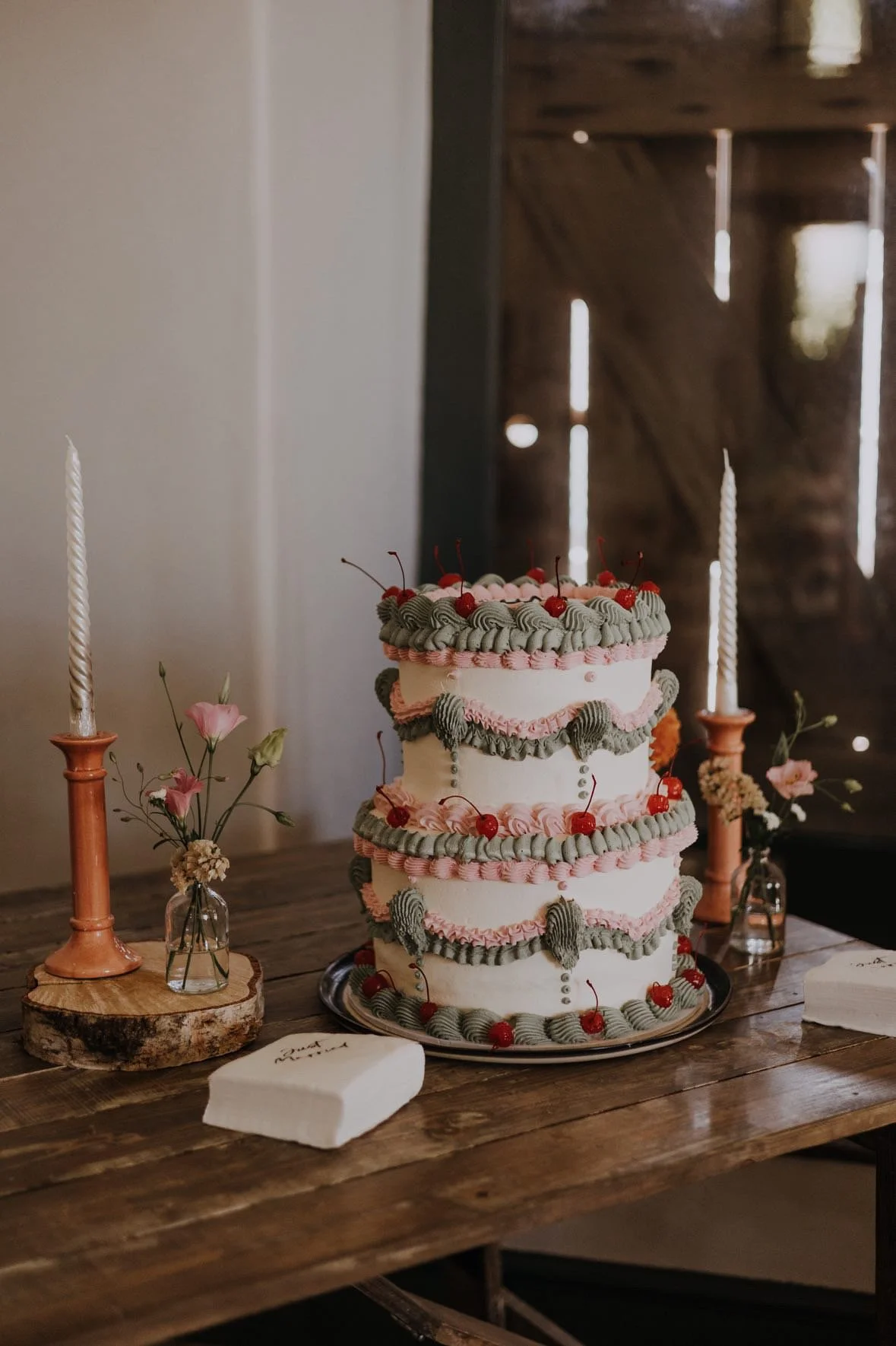 A multi-layered Lambeth wedding cake decorated with pink and sage buttercream, topped with cherries, on a wooden table with pink candles and small vases with pink flowers.