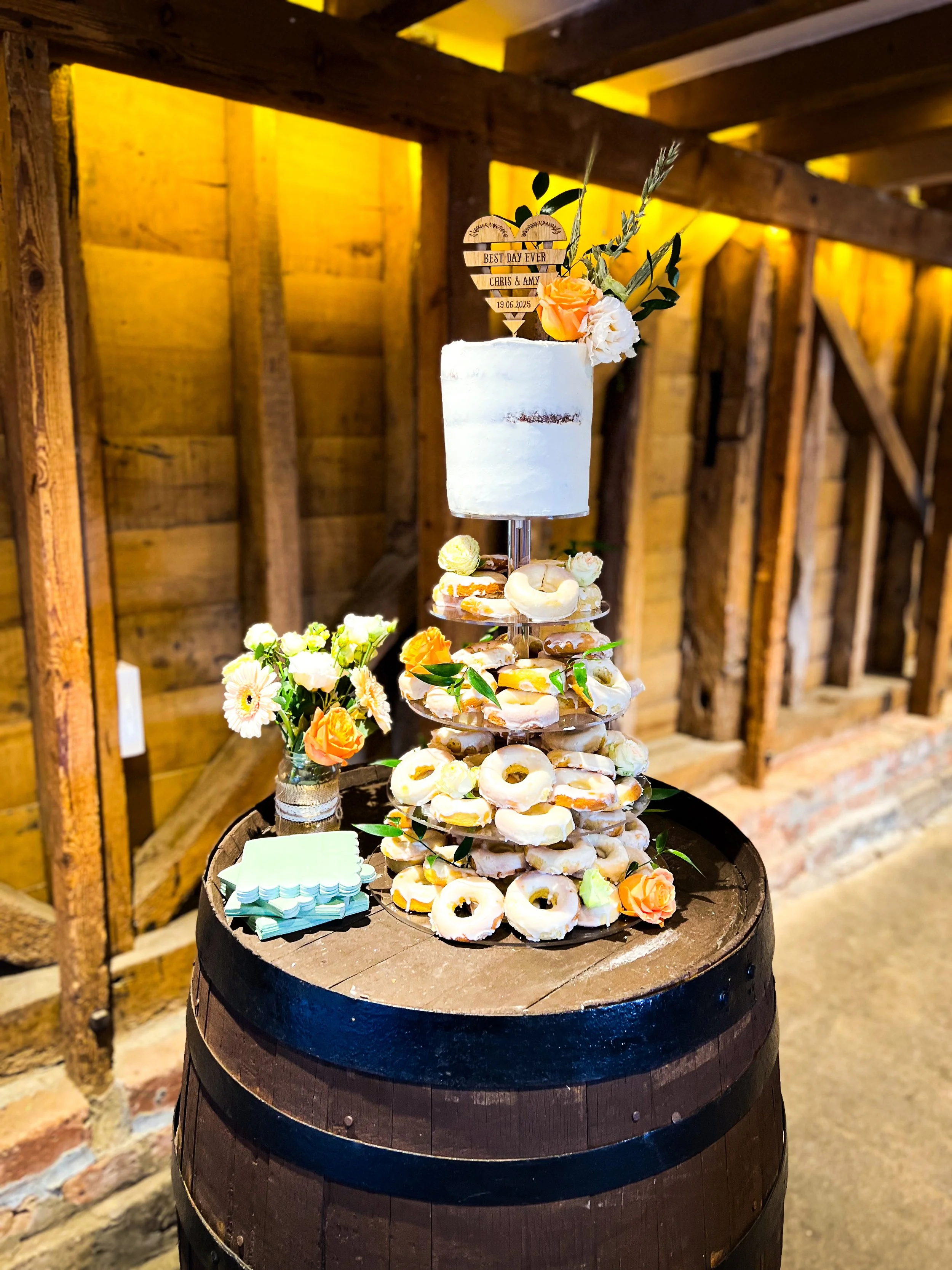 A wedding cake with a white, rustic finish on top of a display of donuts decorated with white icing and citrus slices. The setup is on a wooden barrel, with a small bouquet of flowers and a stack of napkins nearby. The background features warm wooden