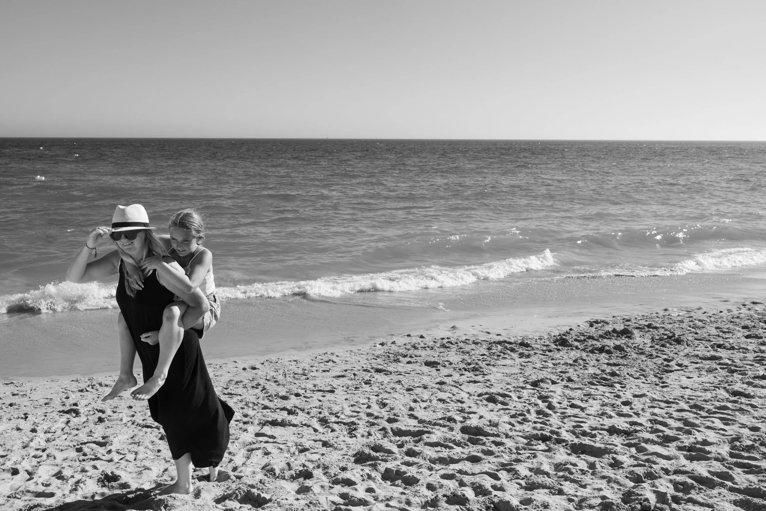 On a beach during a vacation session, Michelle Molnar Photography photographs a mom giving her daughter a piggyback ride.