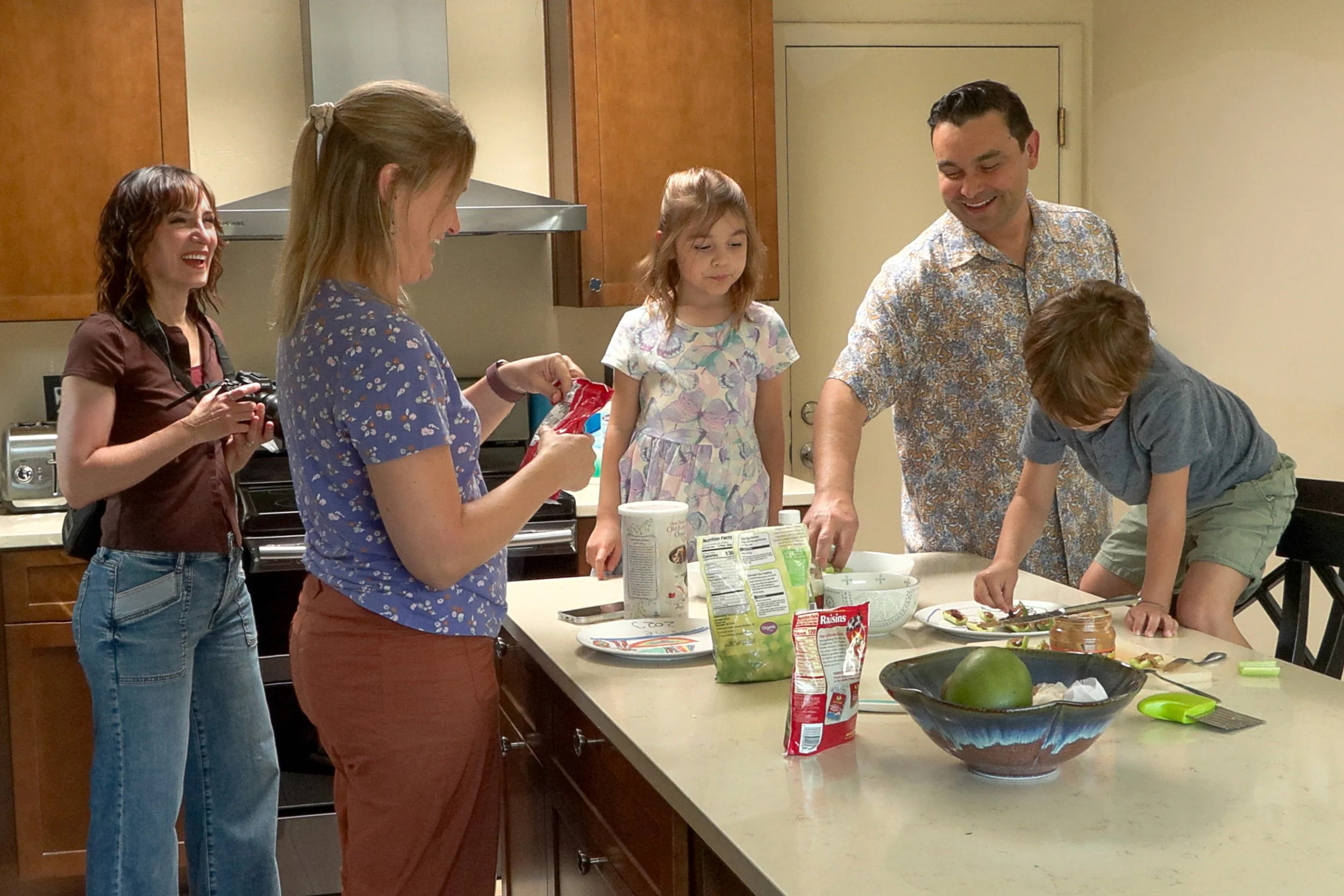 A family gathered around a kitchen island preparing food and baking together. There are two women, one man, and three children, with various snacks and ingredients on the counter. Created by Tucson Family photographer, Michelle Molnar.