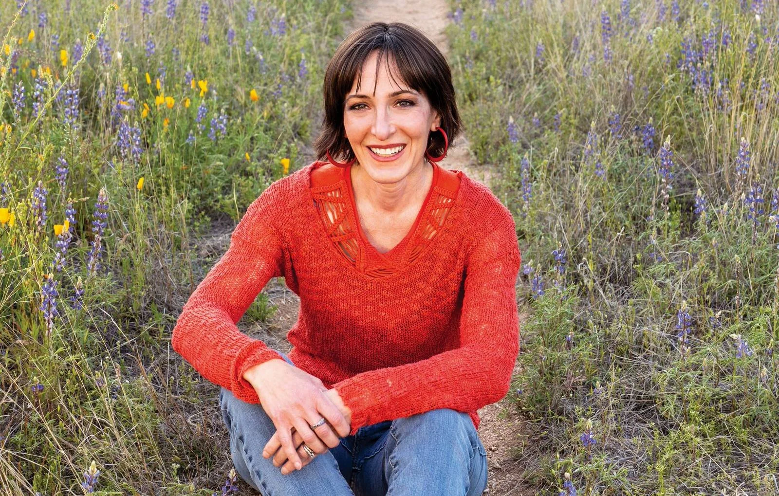 A woman with short brown hair, wearing a red sweater and blue jeans, sitting on a trail in a field of purple and yellow wildflowers, smiling at the camera.