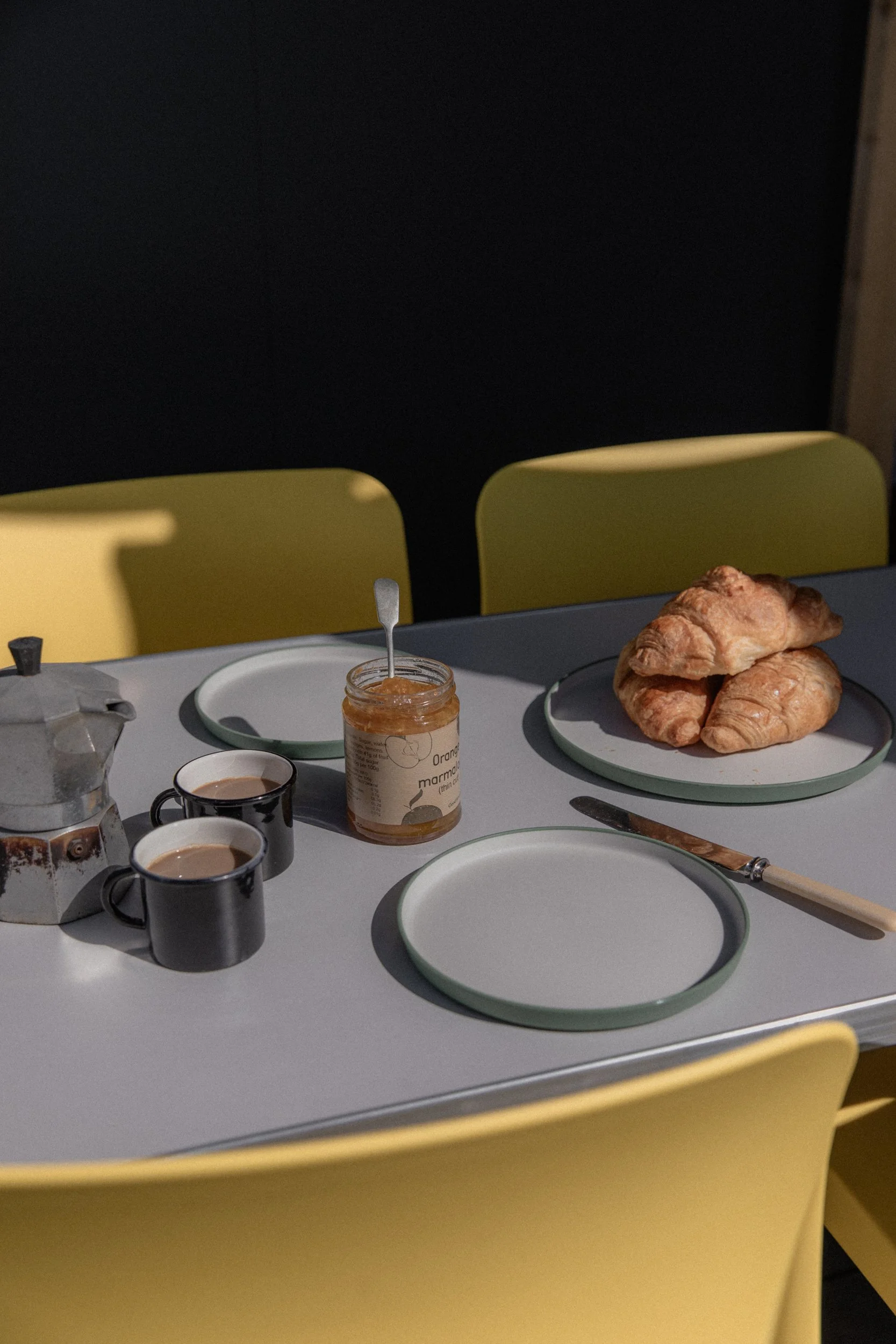 Table set with two black coffee cups, a jar of orange marmalade with a spoon, two croissants on a plate, a butter knife, and a gray teapot, with yellow chairs in the background.